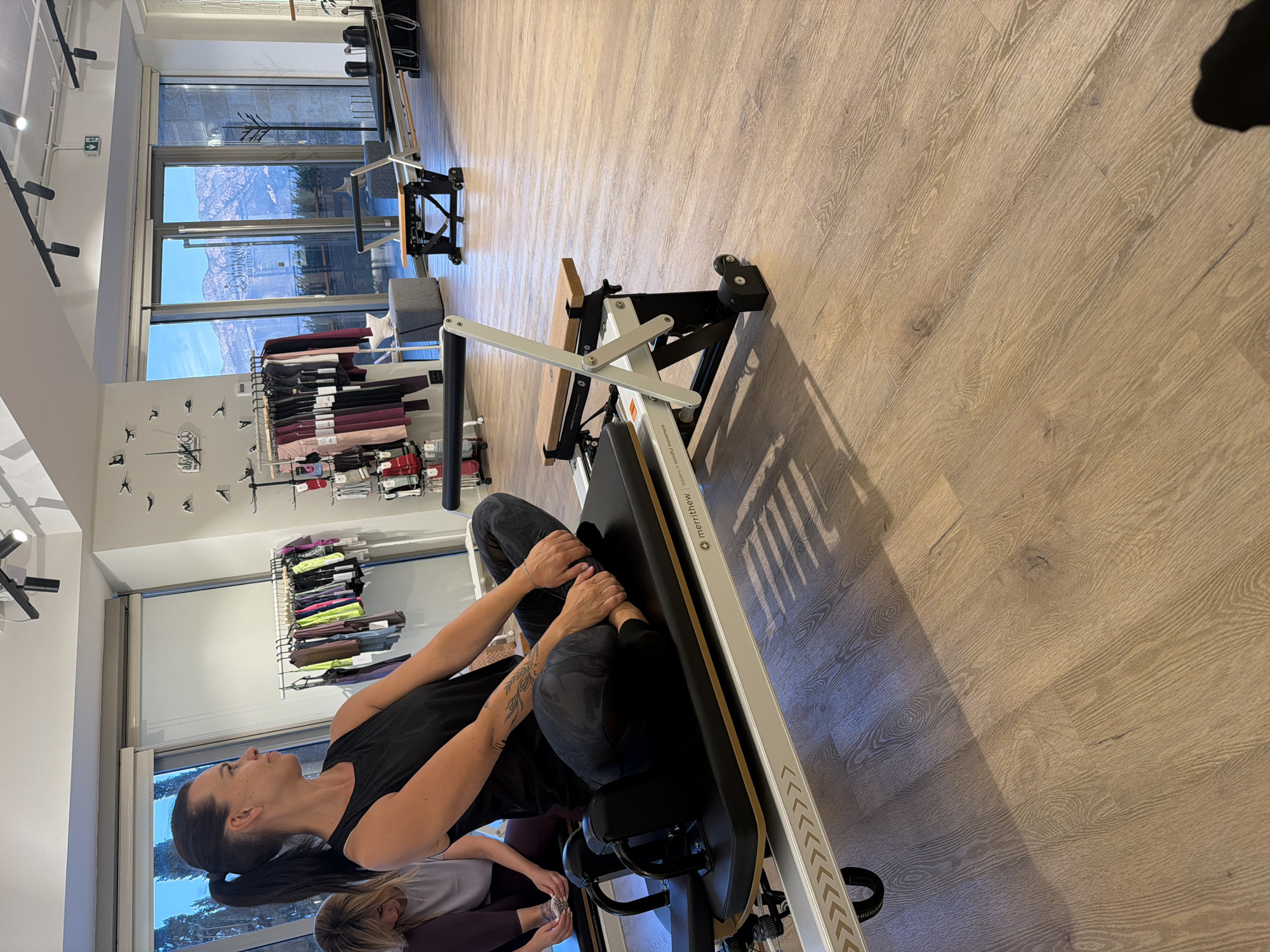 The image depicts an indoor pilates studio, likely located in Boreti, Montenegro, given the scenic mountain and water views visible through the large windows. The primary focus is on a woman with dark hair tied in a ponytail, wearing a black tank top and patterned leggings, seated on a pilates reformer in a cross-legged position. She has a tattoo on her left forearm. To her left, another woman with blonde hair is partially visible, also seated on a reformer. The studio features polished wooden floors and multiple pilates reformers, some of which are positioned in the foreground and midground. Racks of athletic apparel, predominantly leggings and tops in various colors, are displayed against a wall in the background. The overall ambiance suggests a modern and well-equipped fitness facility.