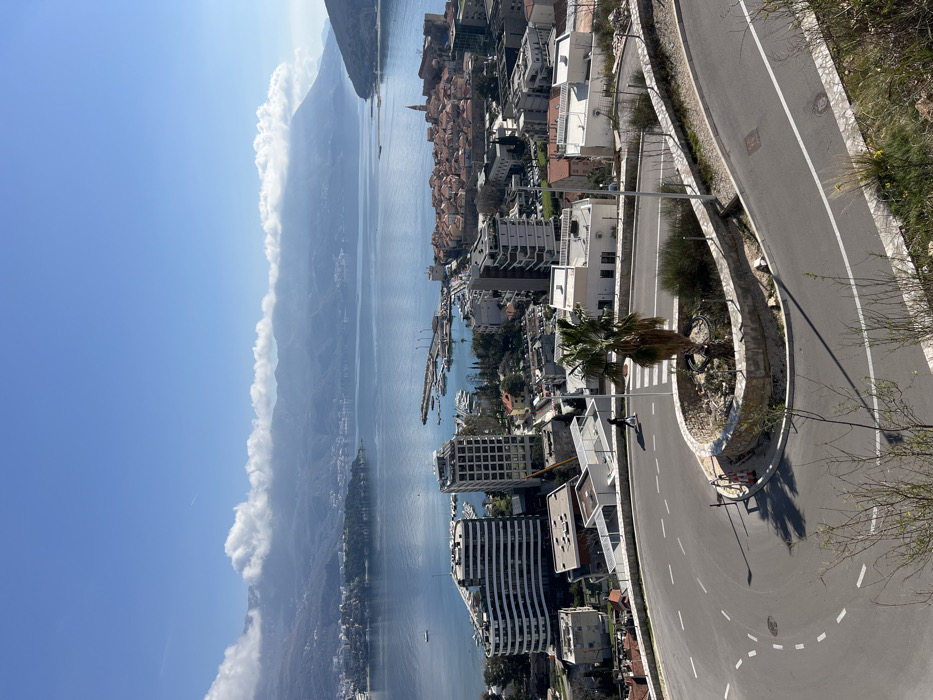 An elevated, panoramic view captures the city of Budva, Montenegro, under clear daytime conditions. In the foreground, a paved two-lane road executes a sharp hairpin turn, delineated by white dashed lane markings. A curvilinear median island within the turn features a single palm tree, sparse vegetation, and rocks. A pedestrian crosswalk is marked on the road's curve. One individual, dressed in dark clothing, walks on the road near the crosswalk, moving towards the left of the frame. Streetlights are visible along the road's edge, and shadows are cast on the pavement.
The midground presents the urban landscape of Budva. To the left, several modern multi-story buildings with distinctive curved facades stand near the coastline. Adjacent to these, a yacht marina is visible in the bay, populated with numerous docked boats. To the right of the marina, the historic Old Town (Stari Grad) is prominent, characterized by its dense cluster of red-tiled roofs, fortified walls, and a visible bell tower. Other residential and commercial buildings are dispersed throughout the urban area.
The background consists of the extensive blue waters of the Adriatic Sea, which form Budva Bay. Large mountains frame the bay on both sides, their peaks partially obscured by a band of white cumulus clouds. An island or peninsula is visible within the bay's waters. The sky above is largely clear and blue, with a faint aircraft contrail present in the upper left quadrant.