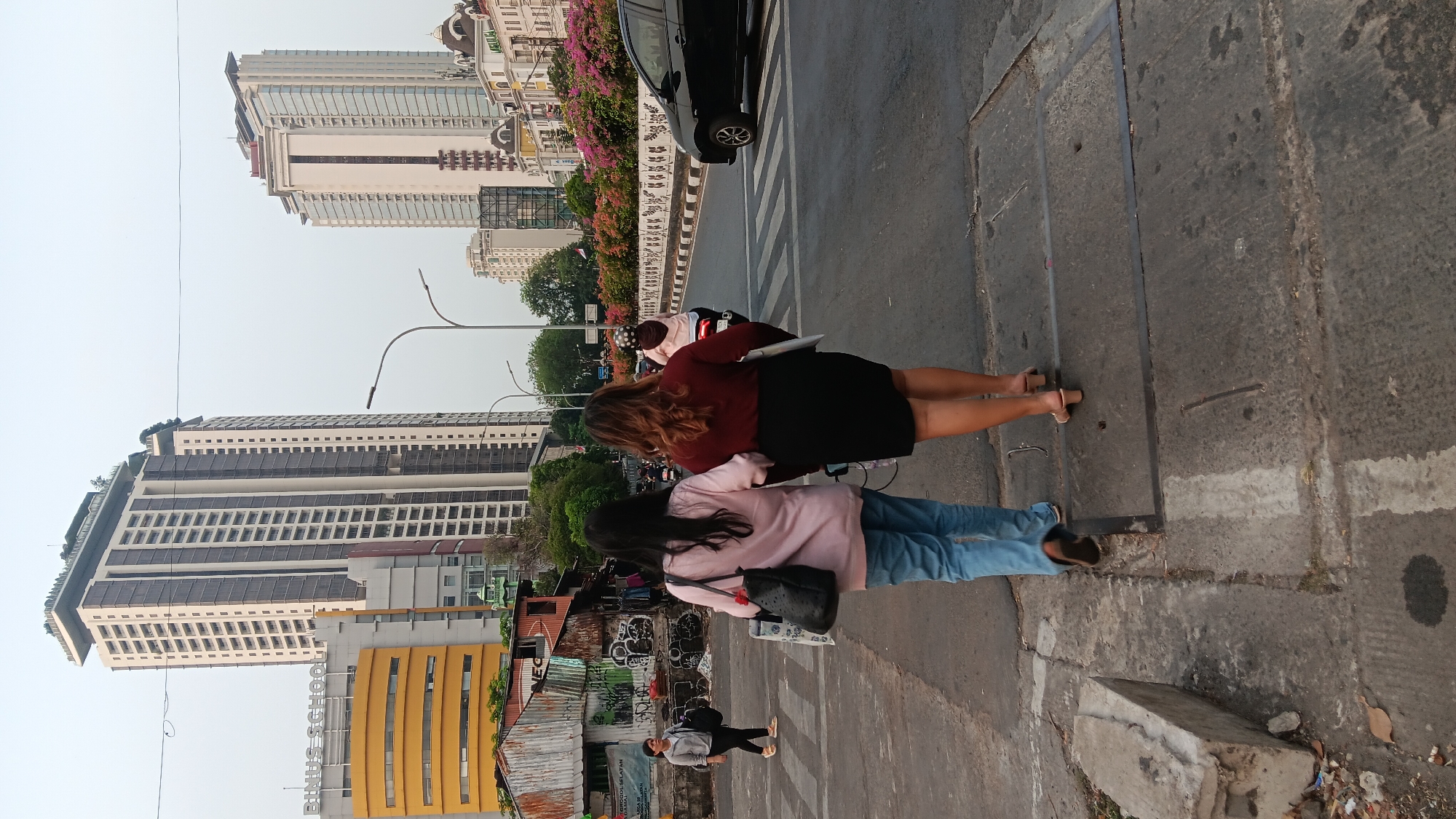 The image depicts a street scene in South Jakarta, Indonesia, likely in the Kebayoran Baru area, known for its modern high-rise buildings and commercial establishments. Two women are walking away from the camera, arm in arm, across a pedestrian crossing on a paved road. The woman on the left wears a light pink long-sleeved shirt and blue jeans, carrying a black tote bag and a white shopping bag. The woman on the right is dressed in a dark red long-sleeved top and a black skirt, with light brown heeled sandals. They are walking towards a row of shops and buildings.
In the background, several tall buildings are visible. The most prominent is a modern, multi-story skyscraper with a white facade and dark windows, featuring green plants on its rooftop. To its left, a yellow building with a curved facade displays the signage "BINUS SCHOOL". Further to the right, another contemporary high-rise building with a glass facade dominates the skyline. The street is lined with trees and flowering bushes, adding color to the urban landscape. A black car is driving on the road to the right of the pedestrian crossing, and a motorcycle with a rider wearing a helmet is also visible on the road. The environment appears to be urban with a mix of modern architecture and some older, more rudimentary structures, suggesting a developing area. The sky is overcast with a light gray hue.