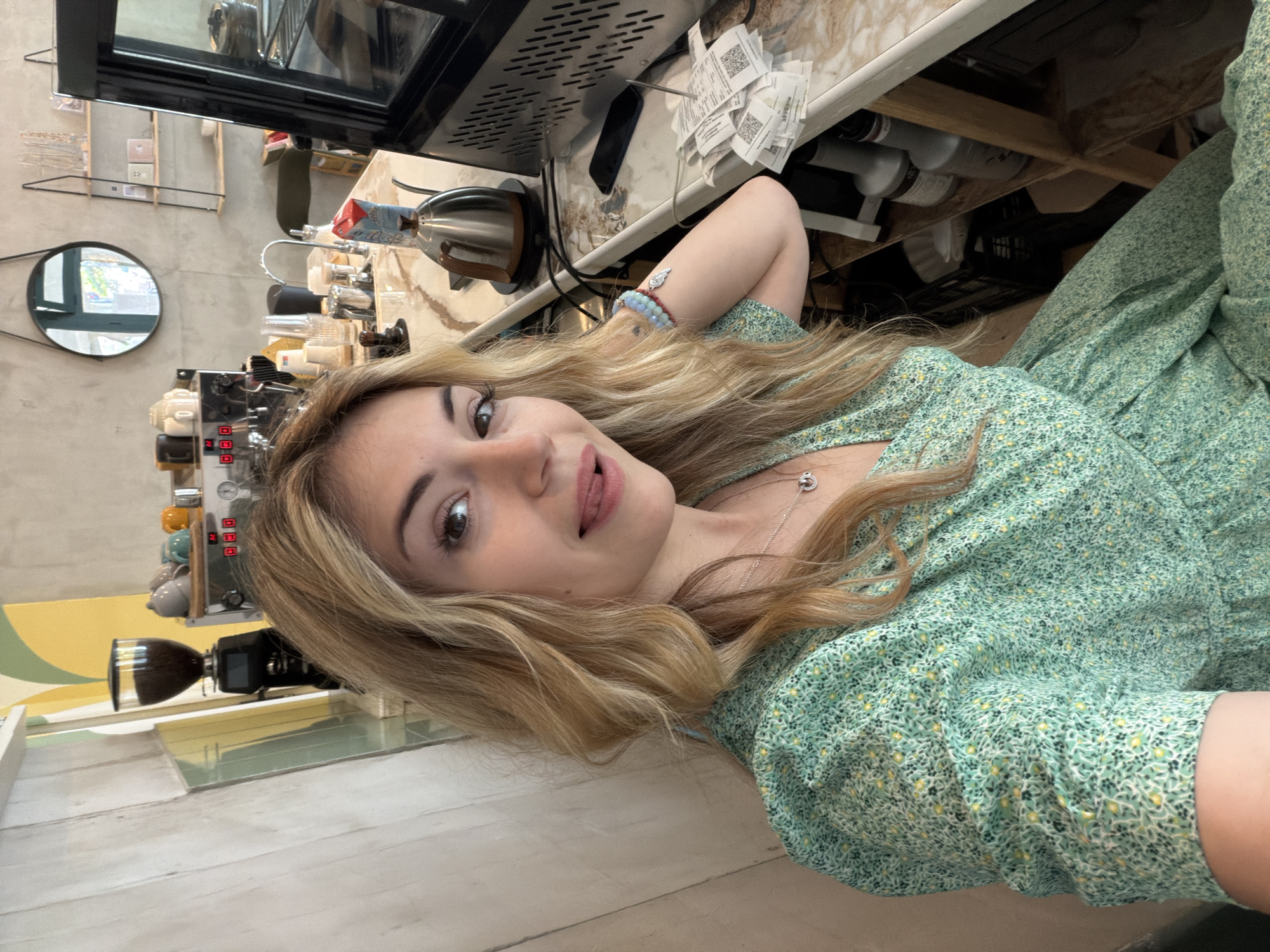 This is a self-portrait (selfie) of a young woman taken indoors, likely within a cafe or coffee shop in Budva, Montenegro. The image is oriented sideways, with the woman's head facing the left of the frame.
The central subject is a young woman with long, wavy blonde hair, fair skin, and light-colored eyes. She is looking directly at the camera with a playful expression, her tongue slightly extended. She is wearing a light green dress adorned with a small, intricate pattern of yellow, white, and dark green leaves or dots. A silver necklace with a circular pendant is visible on her neck, and her right wrist (which is bent behind her head) is adorned with several bracelets, including ones made of blue and white beads and a silver charm.
The background reveals a cafe setting. Behind the woman, there is a counter with various coffee-making equipment, including a silver espresso machine with red indicator lights and buttons, and a coffee grinder with a dark hopper. Various plastic cups and containers are arranged on the counter. Above the counter, a black display case or shelf is visible. A round, black-framed mirror on the back wall reflects an outdoor scene, suggesting daylight outside. The walls appear to be a light gray, possibly concrete, with a decorative yellow and light green painted stripe near the bottom. Beneath the counter, an assortment of bottles and supplies is stored, along with several white paper receipts, some displaying QR codes, stuck to a surface. A blue and red product package is partially visible near the counter. The foreground and floor areas consist of light-colored wooden planks or a similar laminate material.
The overall mood of the image is casual and lighthearted, indicated by the woman's playful expression and pose. There is no specific activity other than the woman taking a selfie. The visible text on the receipts is not legible, but some generic numerical displays and red buttons are seen on the coffee machine.