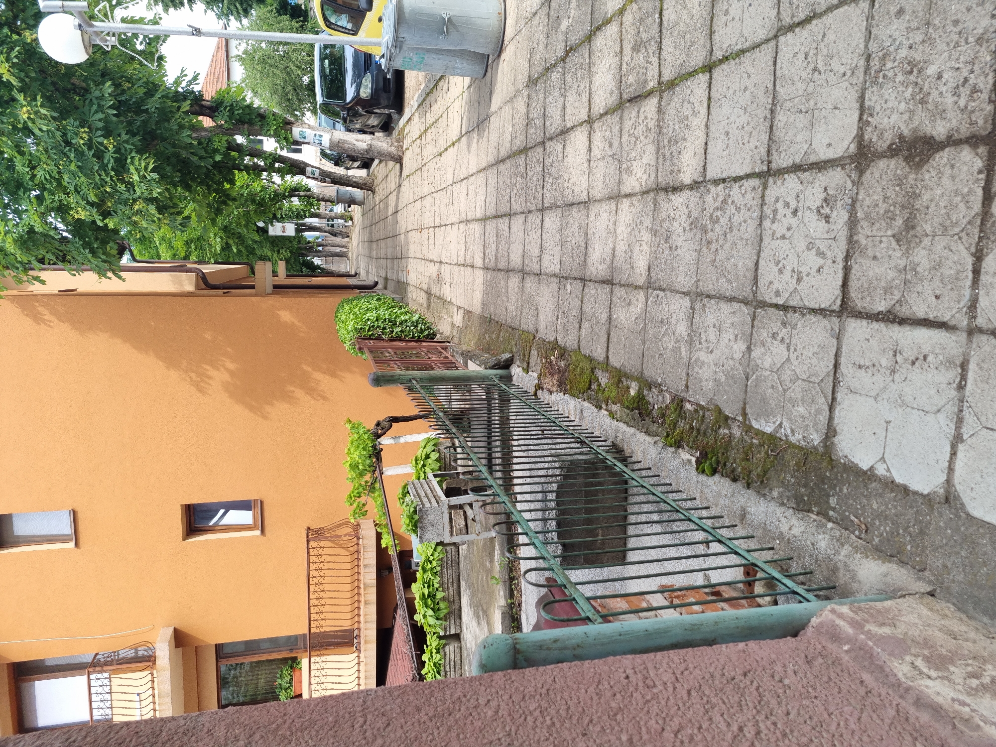 The image depicts a sidewalk in Belogradchik, Bulgaria, adjacent to an orange building. The sidewalk is paved with square concrete tiles and has a slight incline. Trees line the sidewalk on the right side, casting shadows. Several cars are parked along the street next to the sidewalk. Two large, grey trash cans are situated on the sidewalk. A green metal fence runs along the left side of the sidewalk, bordering a residential area with potted plants and a balcony visible on the orange building. The overall scene suggests a typical urban streetscape.