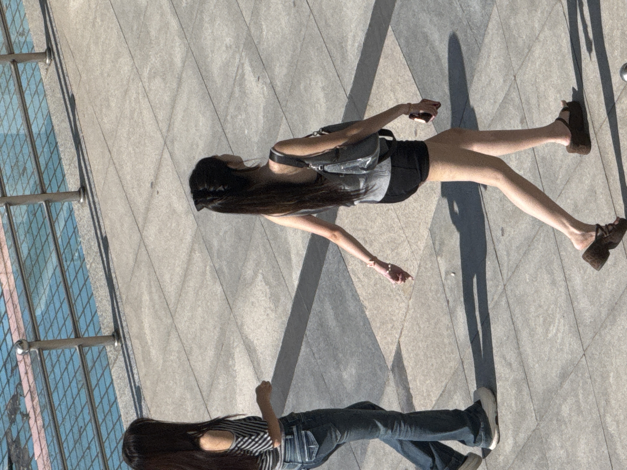 Two young women are walking on a paved outdoor area in Guta, China. The woman on the left is wearing a striped off-the-shoulder shirt and blue jeans. She is walking away from the camera and her hair is dark and long. The woman on the right is wearing a gray tank top and black shorts, carrying a gray shoulder bag and holding a mobile phone. She is walking away from the camera, and her hair is long and dark, tied in a ponytail. The ground is made of grey paving stones. In the background, there is a railing and a body of water with blue tiles. The lighting suggests it is daytime with shadows cast on the ground.