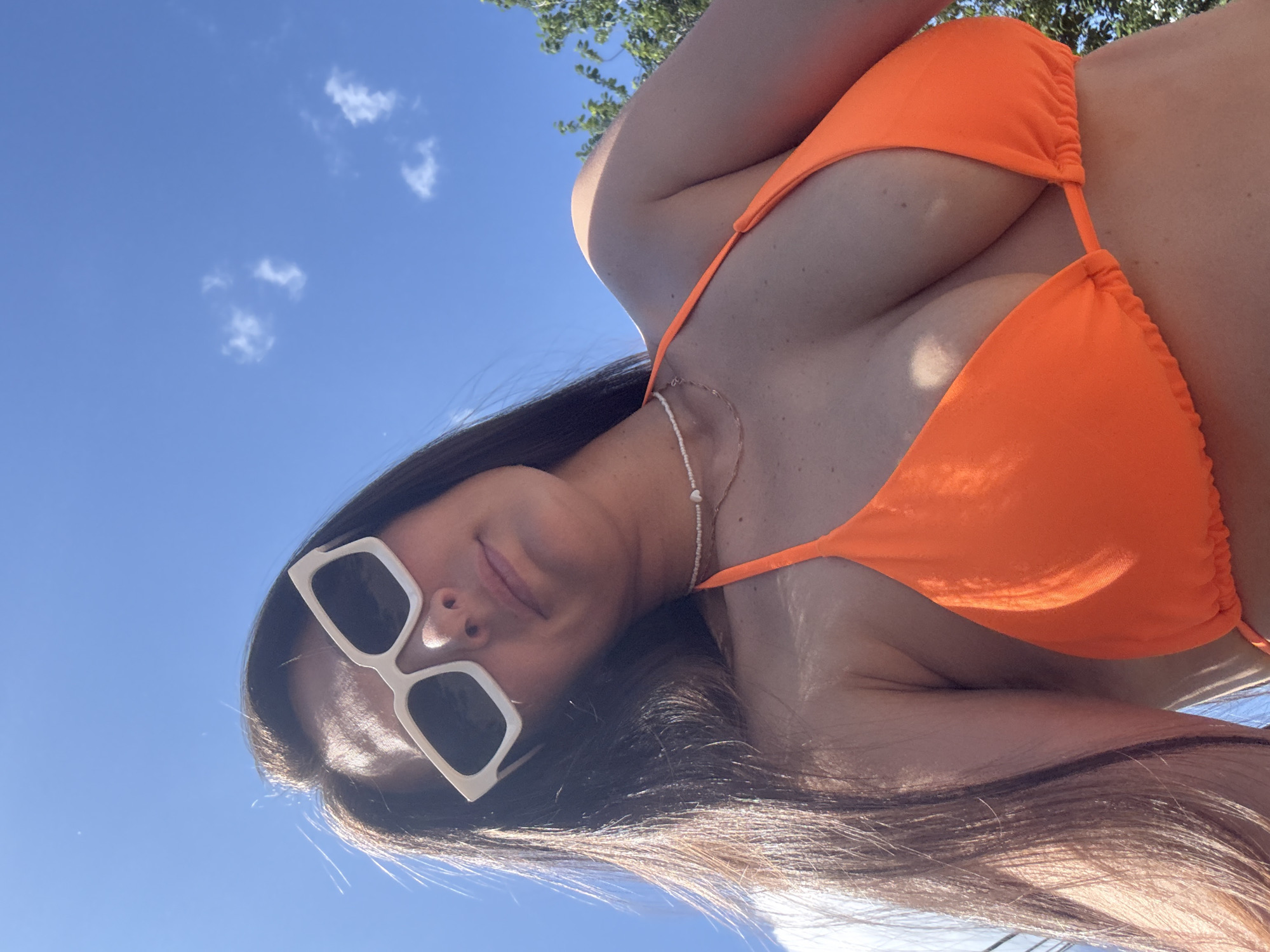 A woman with long, dark hair is wearing an orange bikini and white sunglasses. She is outdoors on a sunny day, with a blue sky and some clouds visible. Trees are also visible in the background. The image appears to be a selfie, taken from a low angle. There is no discernible location in the city of Chaikovsky, Russia.