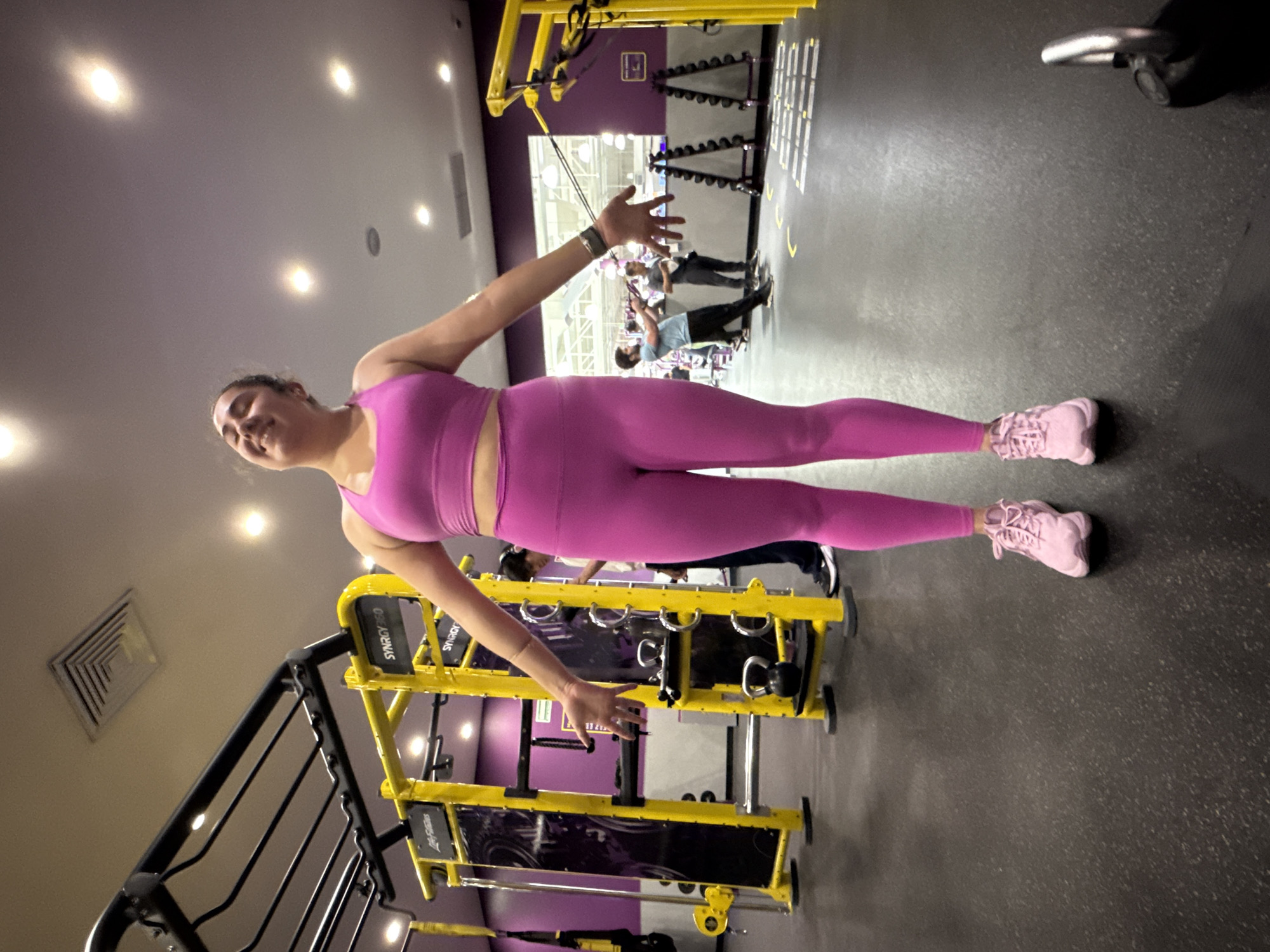 A woman in pink athletic wear (top and leggings) and pink sneakers is in a gym. She stands with arms outstretched. Other people are visible in the background. The gym equipment is predominantly yellow and black. The image may have been taken at a Planet Fitness location, specifically in the city of Puebla, Mexico, due to the brand and color scheme.
