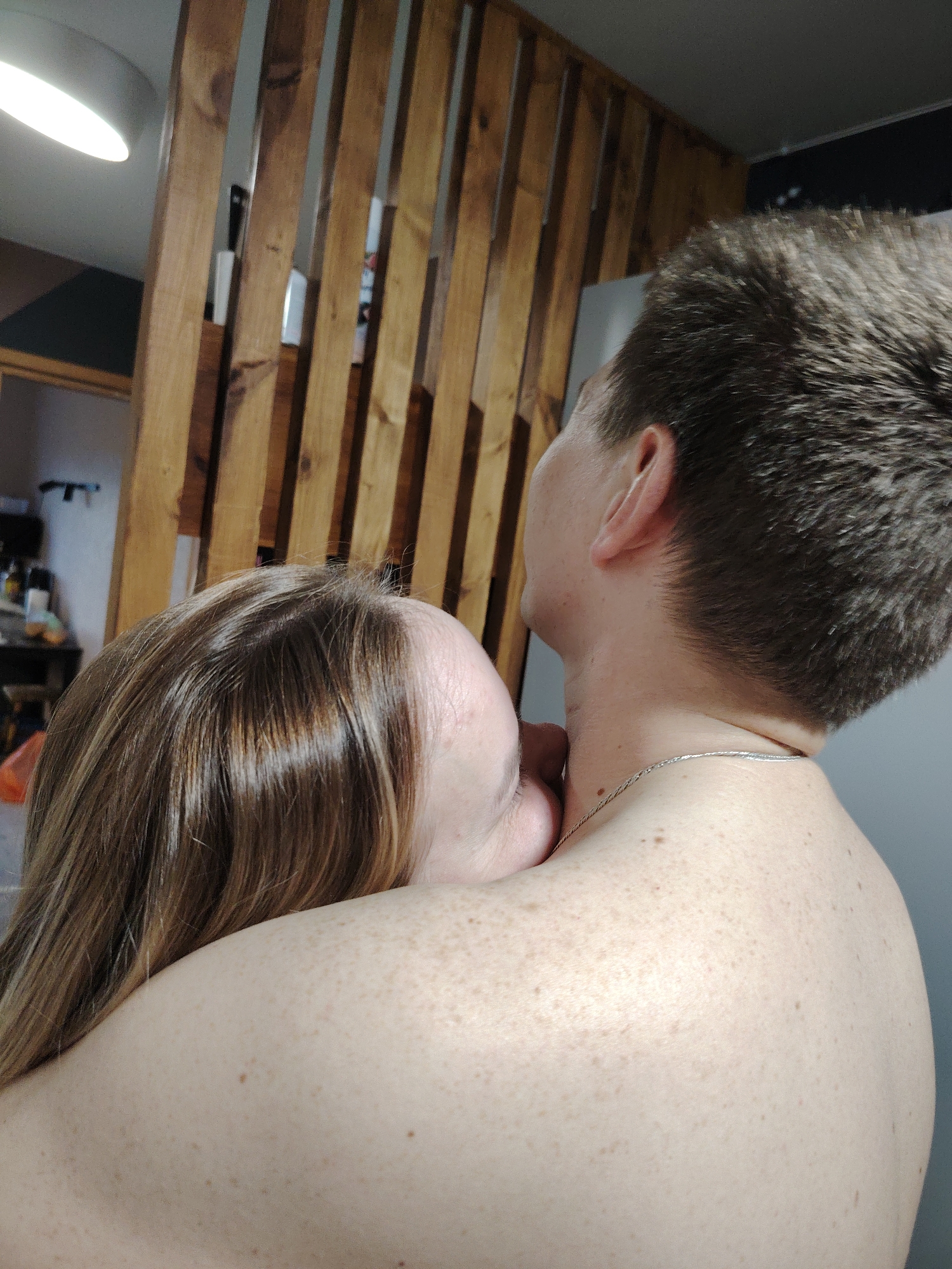A close-up shot depicts a fair-skinned woman with light brown hair embracing a person with short, dark hair and a silver chain necklace. Both individuals are shirtless. The woman's face is partially visible, pressed against the other person's shoulder. The environment features a wooden slat divider and, in the background, a dimly lit room with visible objects. The location is Unknown, Unknown.