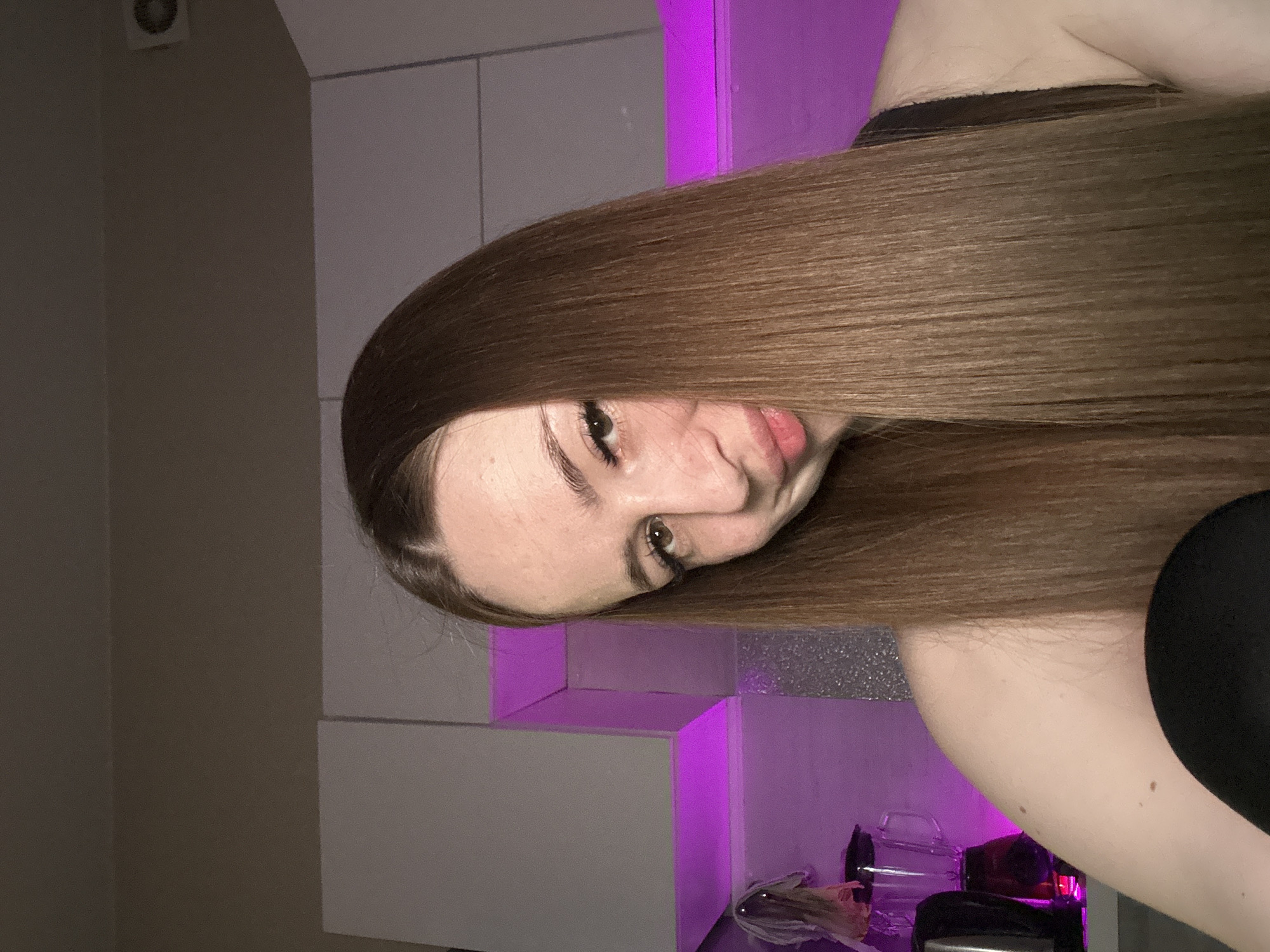 A medium shot shows a person with long, straight brown hair partially obscuring their face. Their hair falls across their face and down the frame, covering most of the background. The person has fair skin, brown eyes, and visible eyelashes. The background appears to be a room with a shelf or ledge, illuminated with a soft purple light. Various small objects are visible on the shelf, but they are not clearly identifiable. The setting appears to be indoors, possibly a personal living space, in a city of Unknown, Unknown.