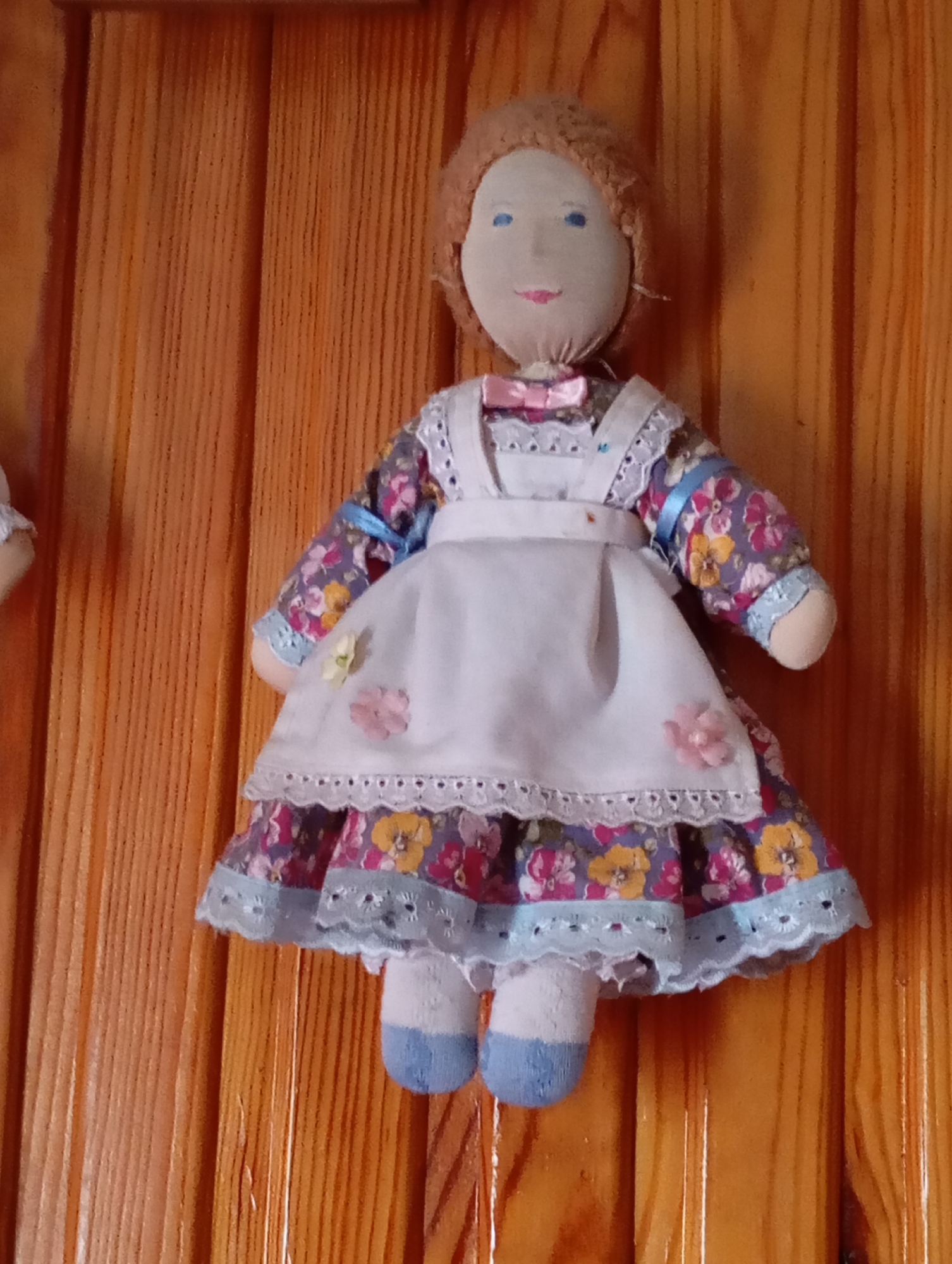 This is a medium-shot, eye-level, indoor photograph of a handmade cloth doll. The doll is dressed in a white apron over a floral-print dress with a purple background and pink, yellow, and blue flowers. The doll has blue eyes, pink lips, and light brown yarn hair. A pink bow is at its neck. The doll's legs are covered in white stockings with blue feet. The doll is attached to a wooden surface, likely a wall, with visible vertical wood grain. The scene is static, with no apparent action or interaction. The location in Льниха, Russia is not visually evident from the image.