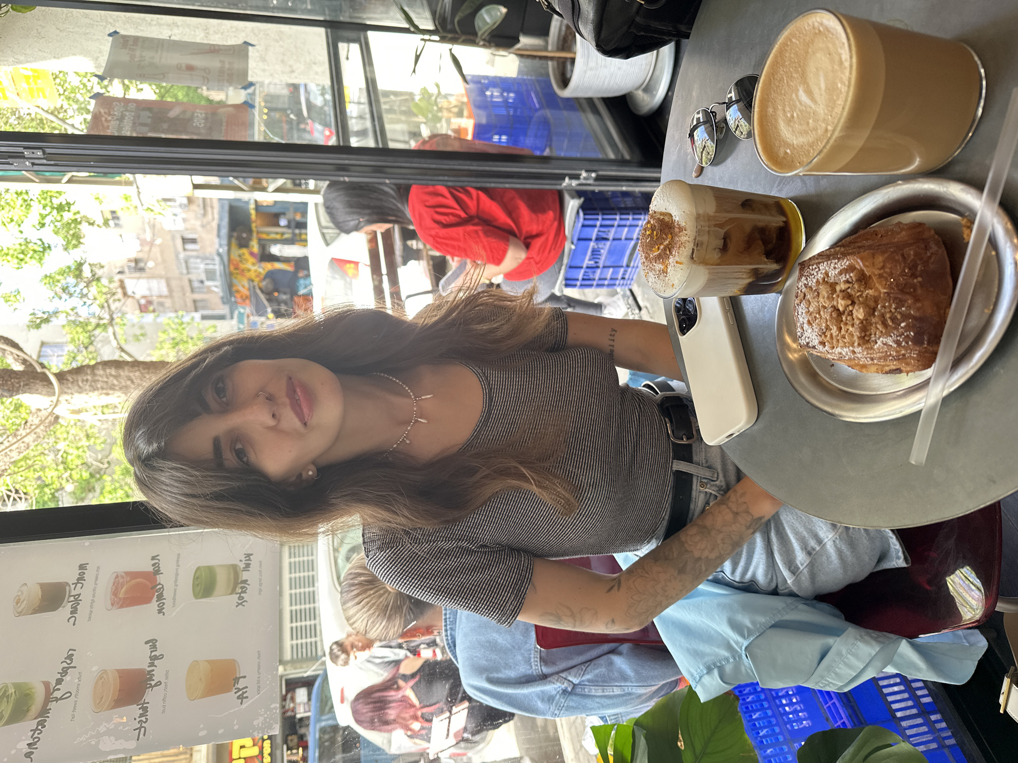 A woman is sitting at a cafe table. The cafe is located in Tel Aviv, Israel, based on signs that are visible in the image. She has a pastry on a plate in front of her along with two drinks and a phone on the table. Her skin is tanned. Her hair is medium brown. She wears a striped t-shirt. She wears a silver necklace and has tattoos on her left arm. The table she's sitting at has a grey top and a red base. In the background, other people are walking in front of the cafe. There are blue plastic crates and plants near the table. A black purse is placed on a shelf behind the woman's head.