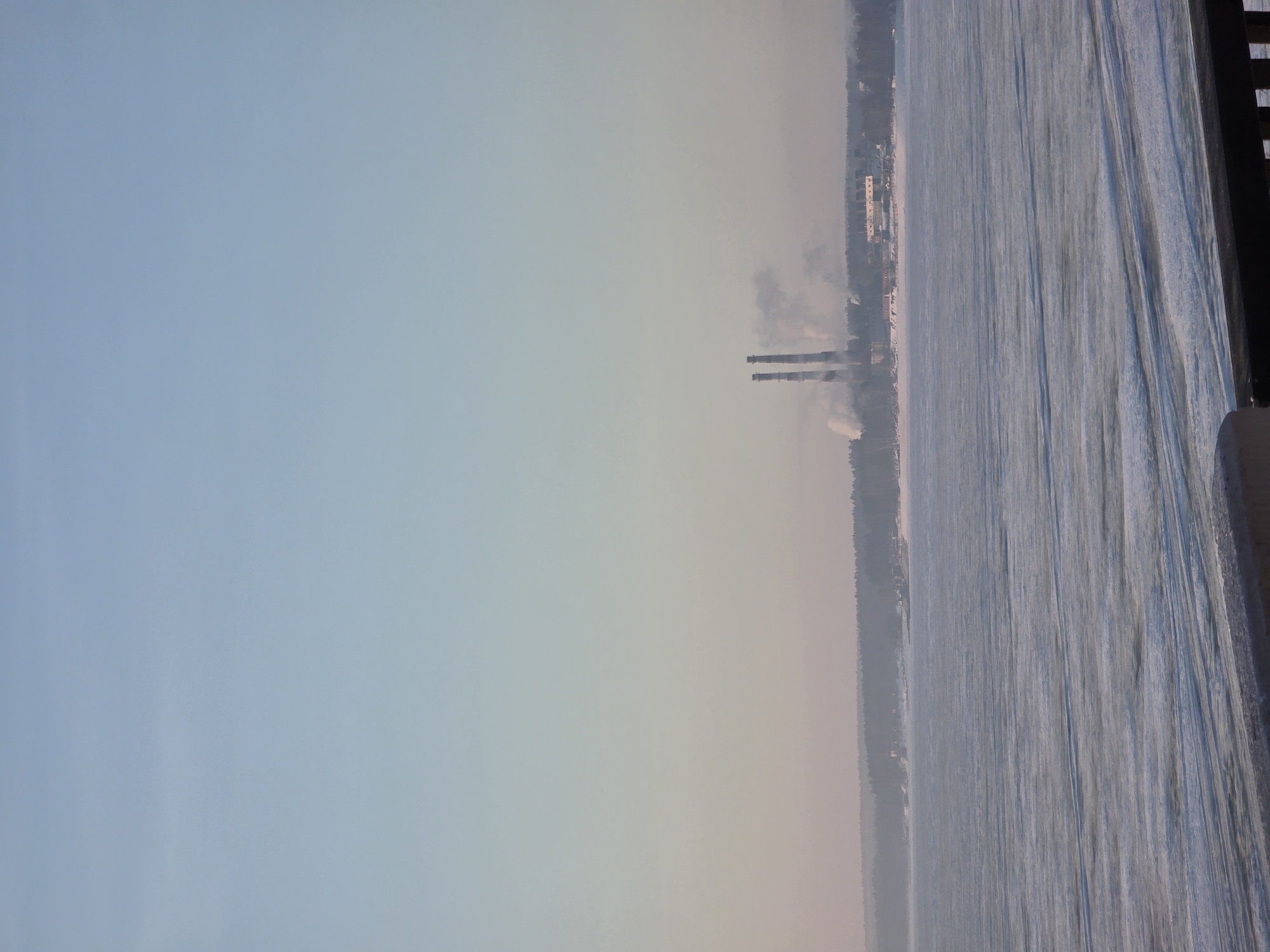 The image captures a wintry scene, likely taken from an elevated position such as a bridge or a high vantage point. The body of water, likely a river or large lake, is mostly frozen over, with visible patterns and textures on the ice surface. In the distance, along the opposite shoreline, there's an industrial complex, recognizable by the presence of two tall smokestacks emitting plumes of smoke. A settlement with some multistoried structures is also visible. The sky is a gradient of blues and grays, indicating overcast or hazy conditions. The city of Chaikovsky, Russia, is likely where this image was captured.