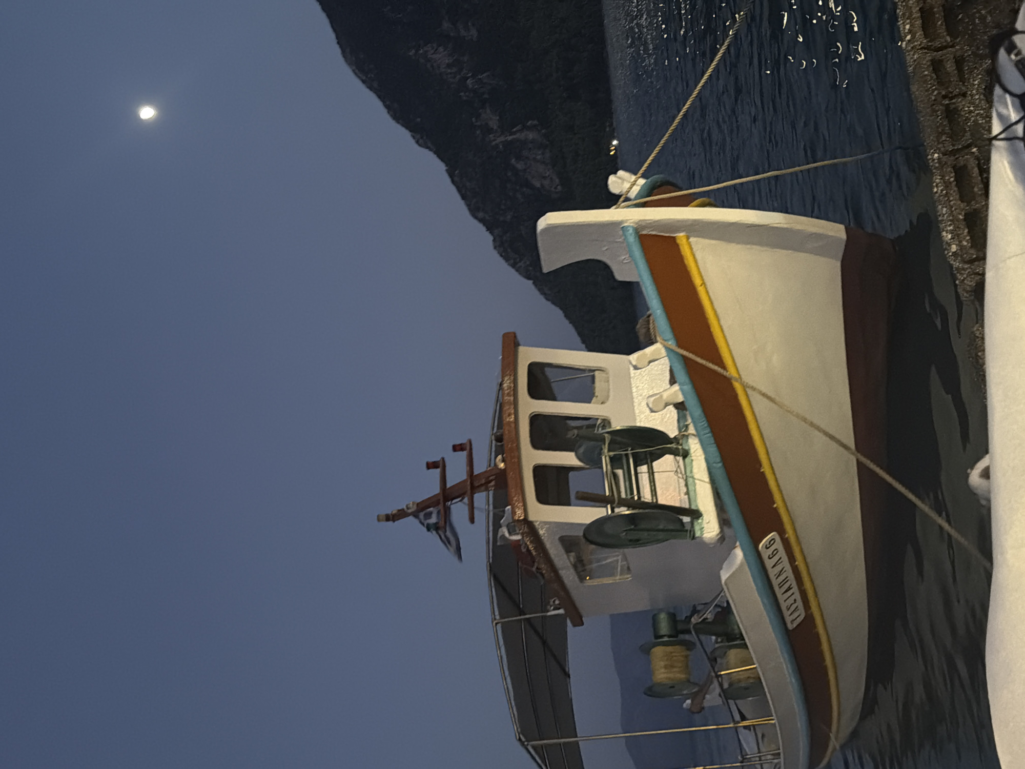 An anchored fishing boat, named "TAZI ANNA 66" with Greek lettering, floats in the dark blue waters near a stone quay. The boat is white with a reddish-brown and blue stripe along its hull, and a yellow accent line. It has a covered canopy and visible fishing equipment, including reels. In the background, a dark, tree-covered mountain rises steeply from the water. The sky is a deep twilight blue, with a bright full moon visible in the upper right quadrant. The scene appears to be in a harbor or bay, likely within the Municipal Unit of Alyzia, Greece, given the context of Greek lettering on the boat and the mountainous coastal landscape. The water reflects the moonlight in shimmering patterns.