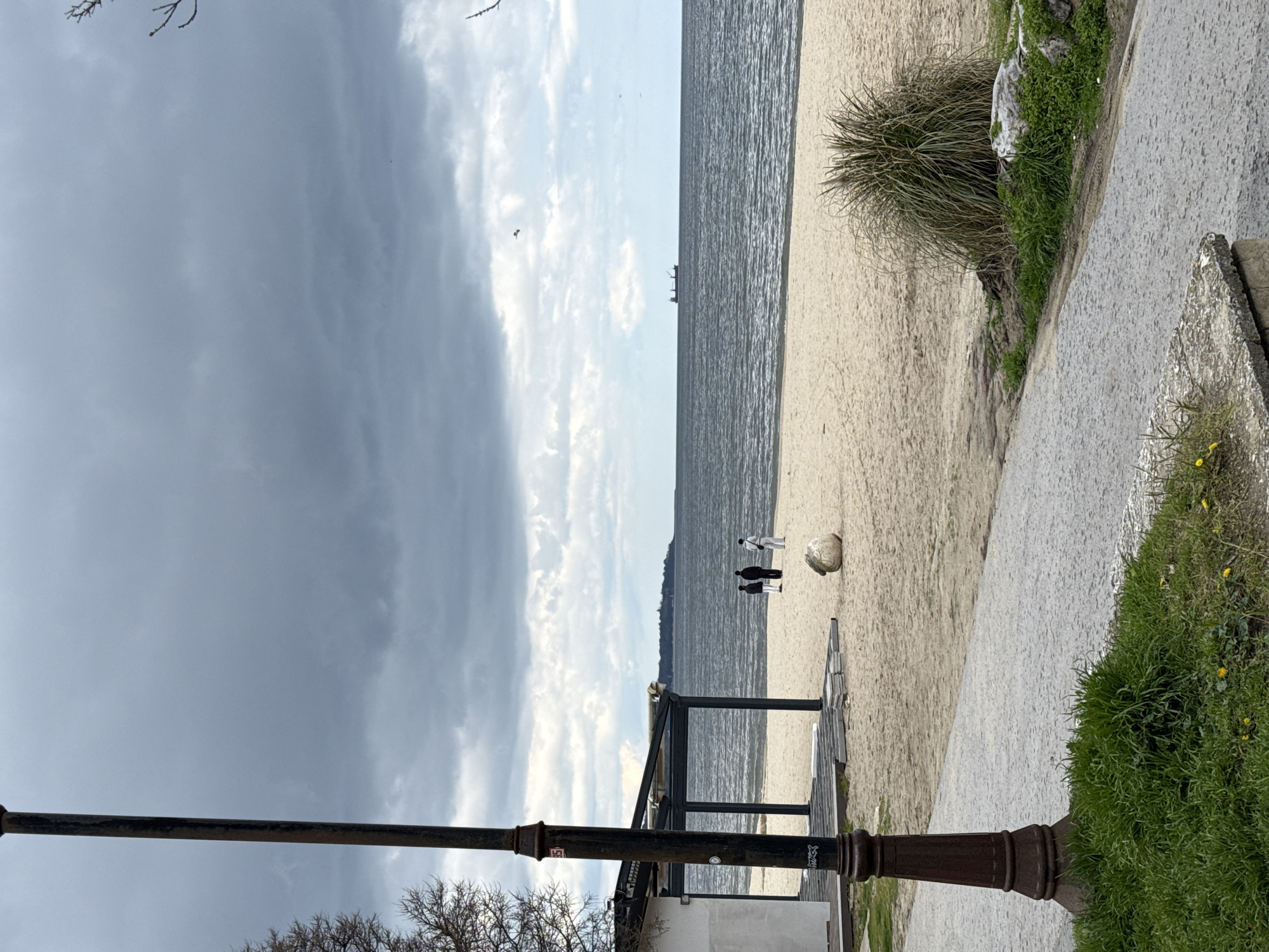 The scene depicts a beach in Varna, Bulgaria, under a sky with dark storm clouds on the left transitioning to partly cloudy skies over the water. Two people are standing on the beach near the water's edge, along with a white soccer ball on the beach. There is a metal structure resembling a partially covered seating area on the beach. A lamppost is positioned on the edge of a paved walkway adjacent to the beach, with vegetation including grass and bushes along the walkway. The sea stretches into the distance with a ship visible on the horizon.