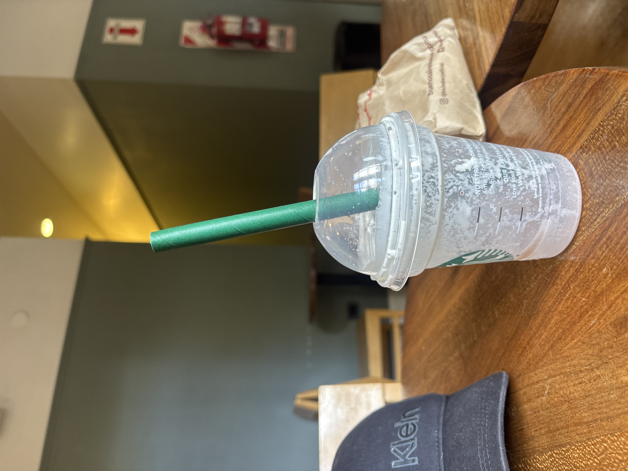 The image shows an empty Starbucks cup with a green straw resting on a wooden table. The cup is transparent and has the Starbucks logo visible. Next to the cup is a small, light brown paper bag. In the background, there is a green wall with a sign displaying an arrow and a sign indicating the presence of a fire extinguisher. Also, there is a partial view of a black cap with the word "Klen" embroidered. The text on the Starbucks cup mentions "Buenos Aires". The location is therefore likely a Starbucks in Buenos Aires, Argentina.
