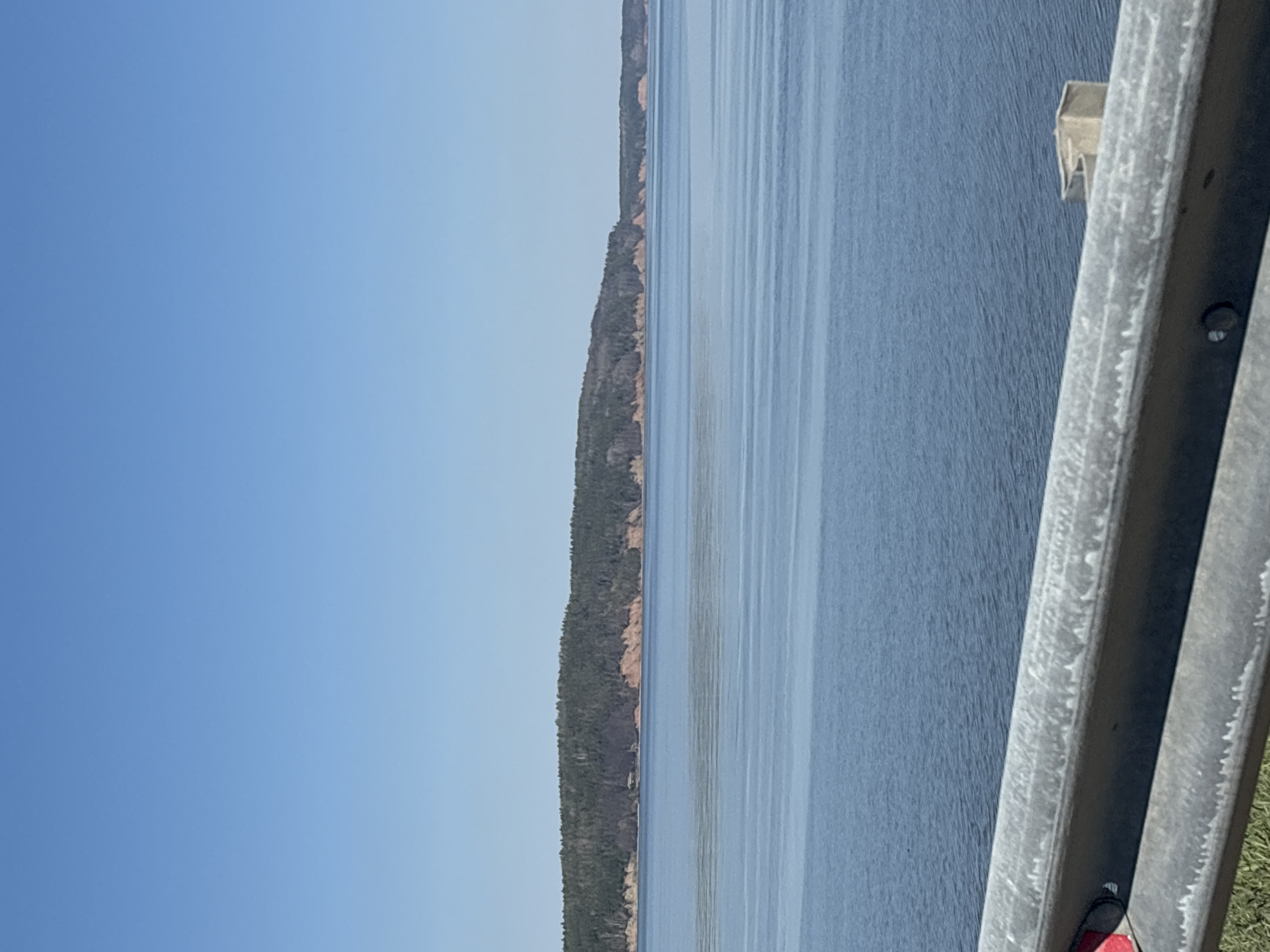 The image shows a body of water, likely a river or a lake, with a heavily wooded shoreline visible on the opposite side. A guardrail is present in the foreground on the right side of the image, suggesting the view is from a road or bridge. The sky is clear and blue. The location is possibly near Chaikovsky, Russia, based on the city mentioned. However, specific details about the location within Chaikovsky, as well as about people, objects, actions or interactions are not visible. The focus is on the landscape view.
