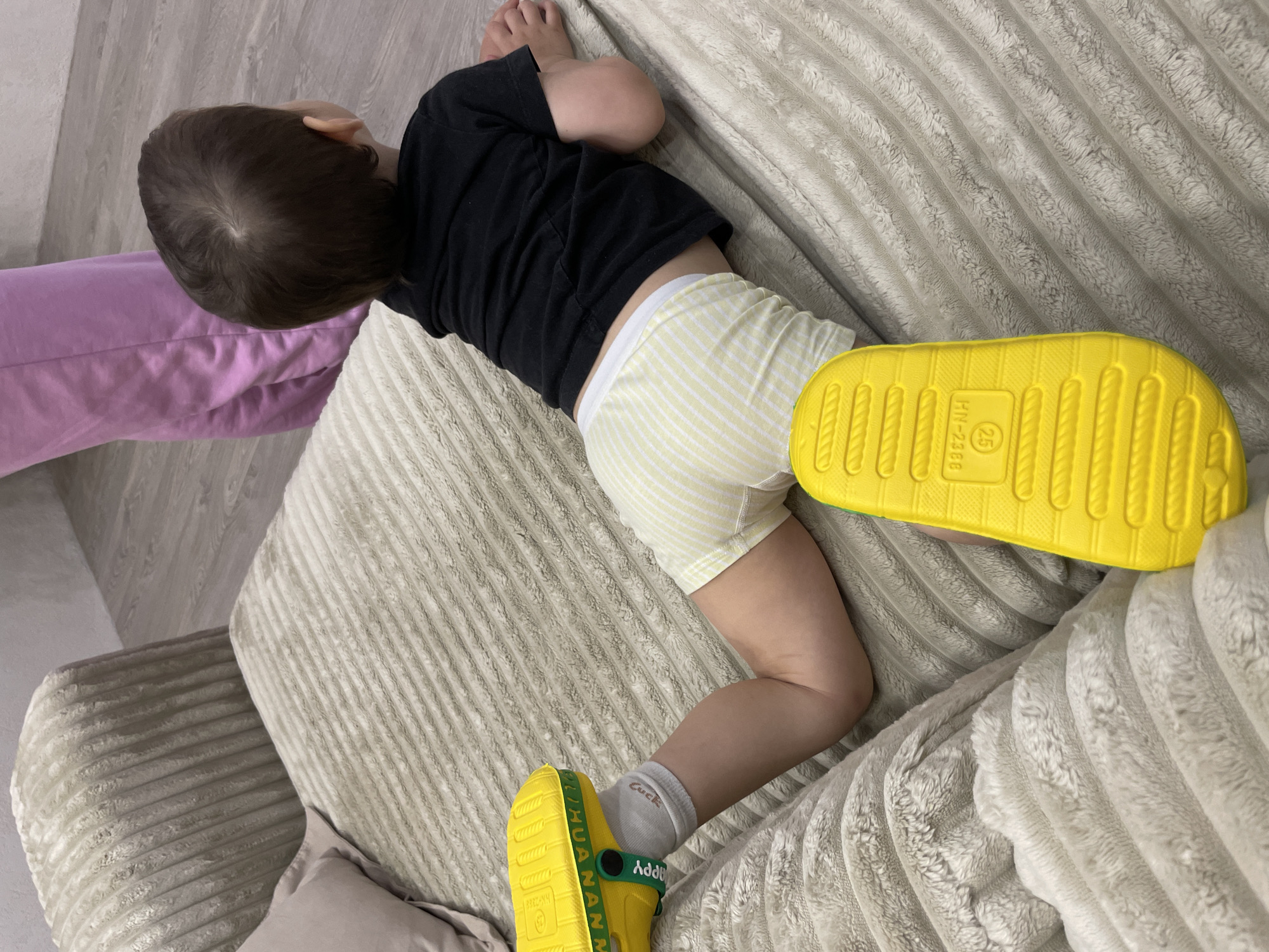 A young child, seen from above and behind, lies on a plush, light-grey ribbed sofa or bed. The child has dark brown hair and is wearing a black short-sleeved t-shirt and yellow and white striped boxer-style shorts. White socks are visible on both feet. On the child's feet are bright yellow slip-on sandals.
The child is positioned with their back to the viewer, head turned slightly to the left, and knees bent. One foot is propped up on the armrest or backrest of the furniture, revealing the textured sole of the yellow sandal. The sole of this sandal has embossed text: "HN-2388" and "25". The other foot rests closer to the child's body, and the green strap of the sandal on this foot shows partially visible text, appearing to read "LIHUANNA" and "Happy" (upside down). A white sock on this foot also has "Luck" written on it.
To the left of the child's head, a piece of purple fabric, possibly a blanket or clothing, is visible. The background consists of a light-colored wooden floor or wall. The setting appears to be indoors, likely a home environment in Izhevsk, Russia. The time of day and weather are not discernible, and the child's expression is not visible.