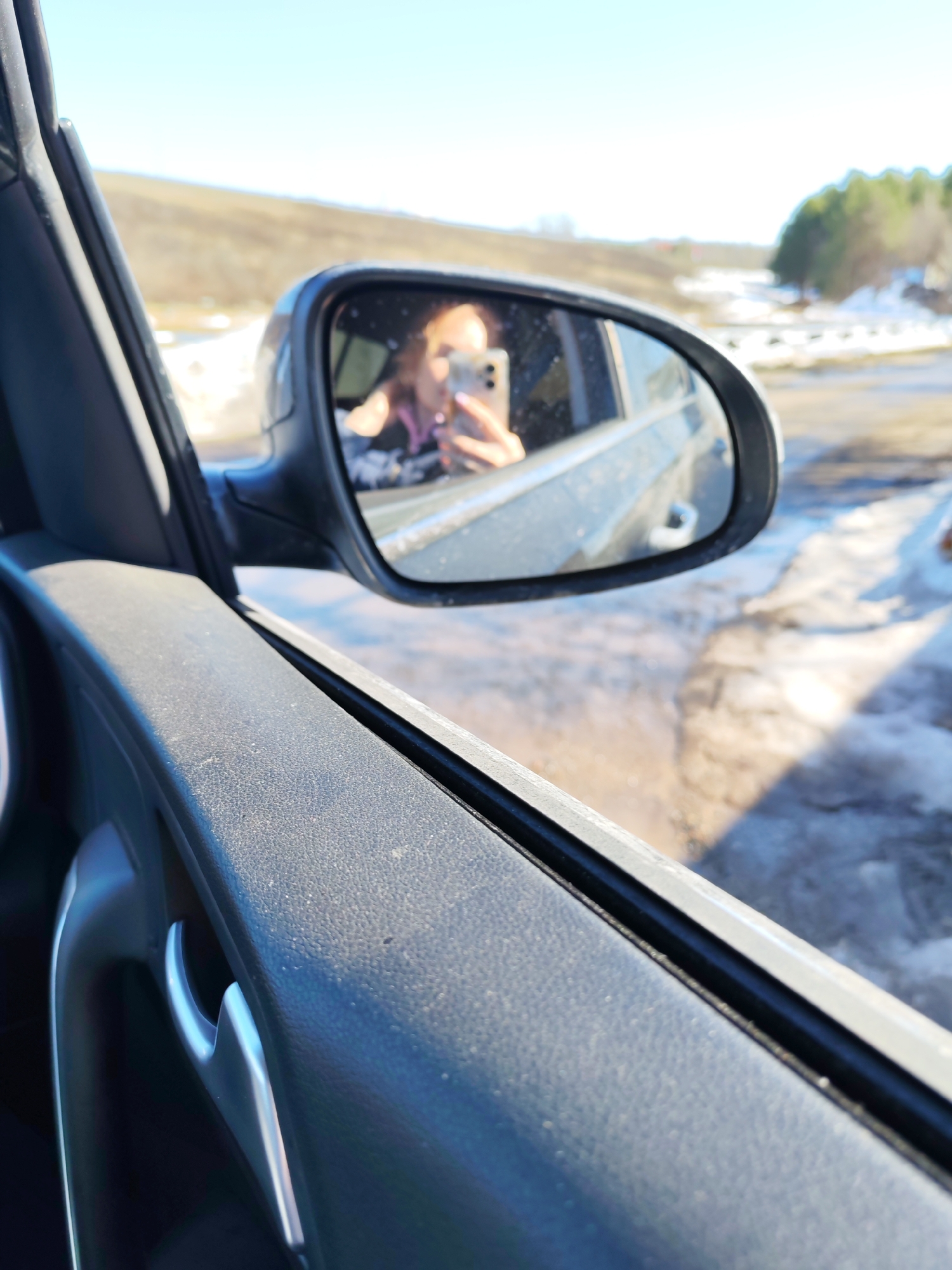 The photograph is taken from inside a car, focusing on the interior door panel and the side mirror. The door panel is textured and dark-colored, with a silver handle visible. The side mirror reflects a woman holding a phone, presumably taking the photo. Outside the car, the landscape is visible, showing patches of snow and a blurred background of trees and possibly a small building. The scene is set in daylight under a clear sky. It can be identified as Chaikovsky, Russia, by the landscape details in the background.