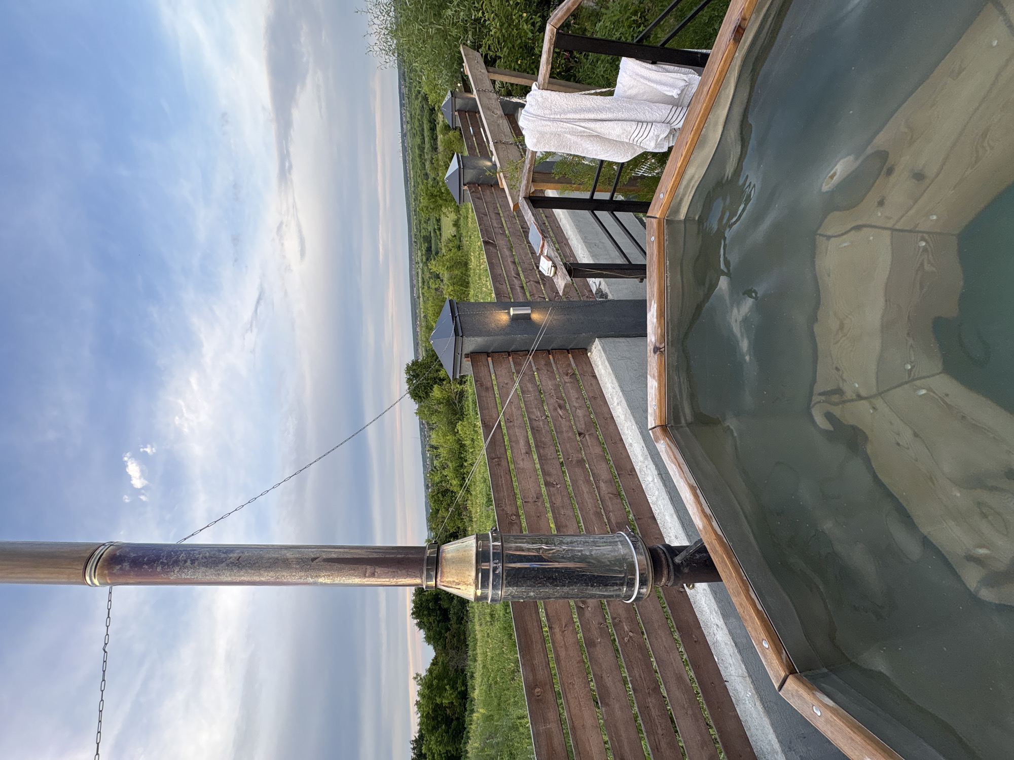 A high-angle, eye-level shot captures a wooden hot tub filled with murky water, positioned on a paved surface. A tall, stainless steel chimney with a tarnished, reddish patina rises from behind the tub. A wooden fence with horizontal planks runs across the midground, separating the tub area from a lush, green landscape with trees and fields that stretches to the horizon under a partly cloudy, pale blue sky. Two modern, dark gray fence posts with small, integrated lights stand along the fence line. A white towel with dark stripes is draped over a black metal railing to the right of the frame. The setting appears to be outdoors, likely in a rural or semi-rural area, potentially a private property given the amenities. The overall scene suggests a relaxing outdoor space with a view.