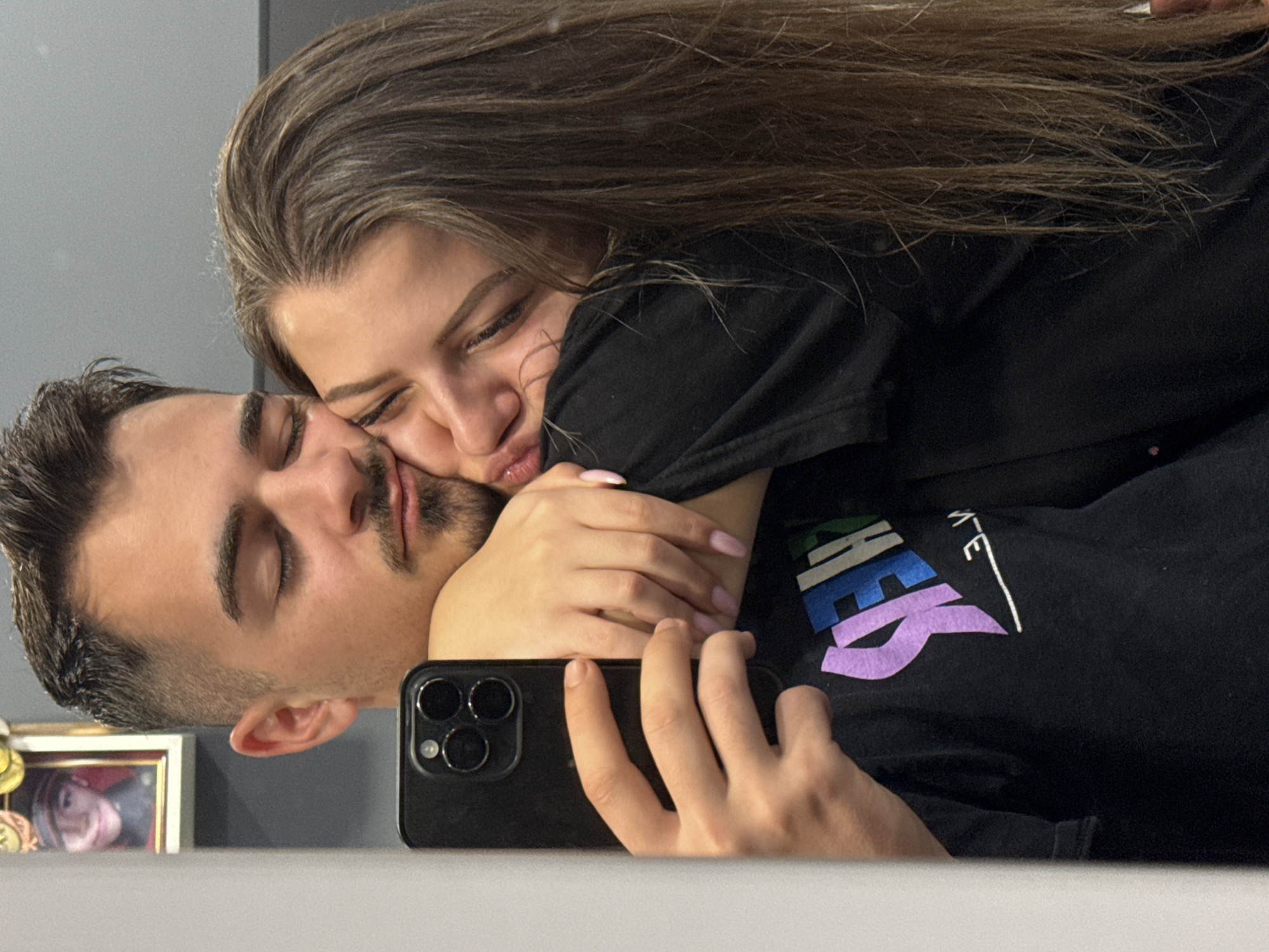 A selfie captures a woman embracing a man closely. The man has short dark hair, a trimmed beard, and closed eyes. The woman has long, light brown hair and her eyes are closed. She is puckering her lips as if to kiss him. Her hands are wrapped around him. They both wear dark-colored t-shirts. The woman's t-shirt has a design on it. The phone is visible in the foreground. In the background, a framed picture sits on a surface. The environment appears to be indoors, likely a residential setting. There are no elements suggesting the city of Sofia, Bulgaria.