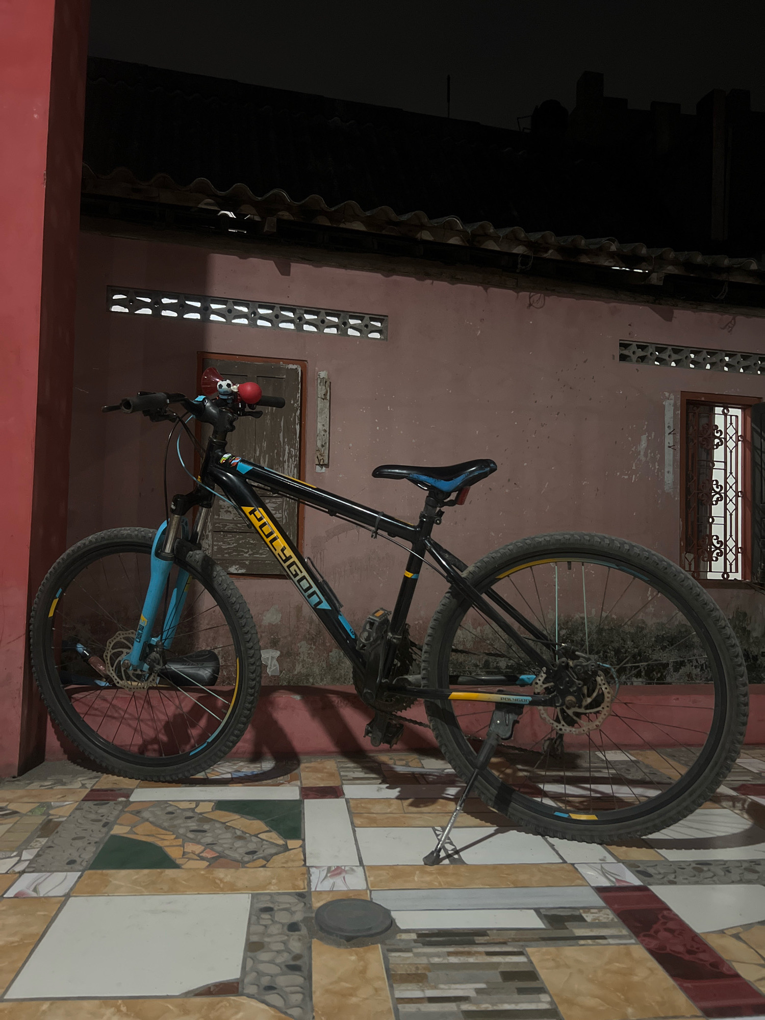 A black Polygon bicycle with blue accents is parked on a tiled patio at night. The bicycle has a kickstand, disc brakes, and a red bell on the handlebars. The wall behind the bicycle is pink, with barred windows and ventilation slits. A red structural column is to the left of the bicycle. The roof of the building is visible above the wall. The ground is covered in a mosaic tile pattern of various colors and shapes. The overall scene appears to be an outdoor area in front of a residential building in Tlogoadi, Indonesia.