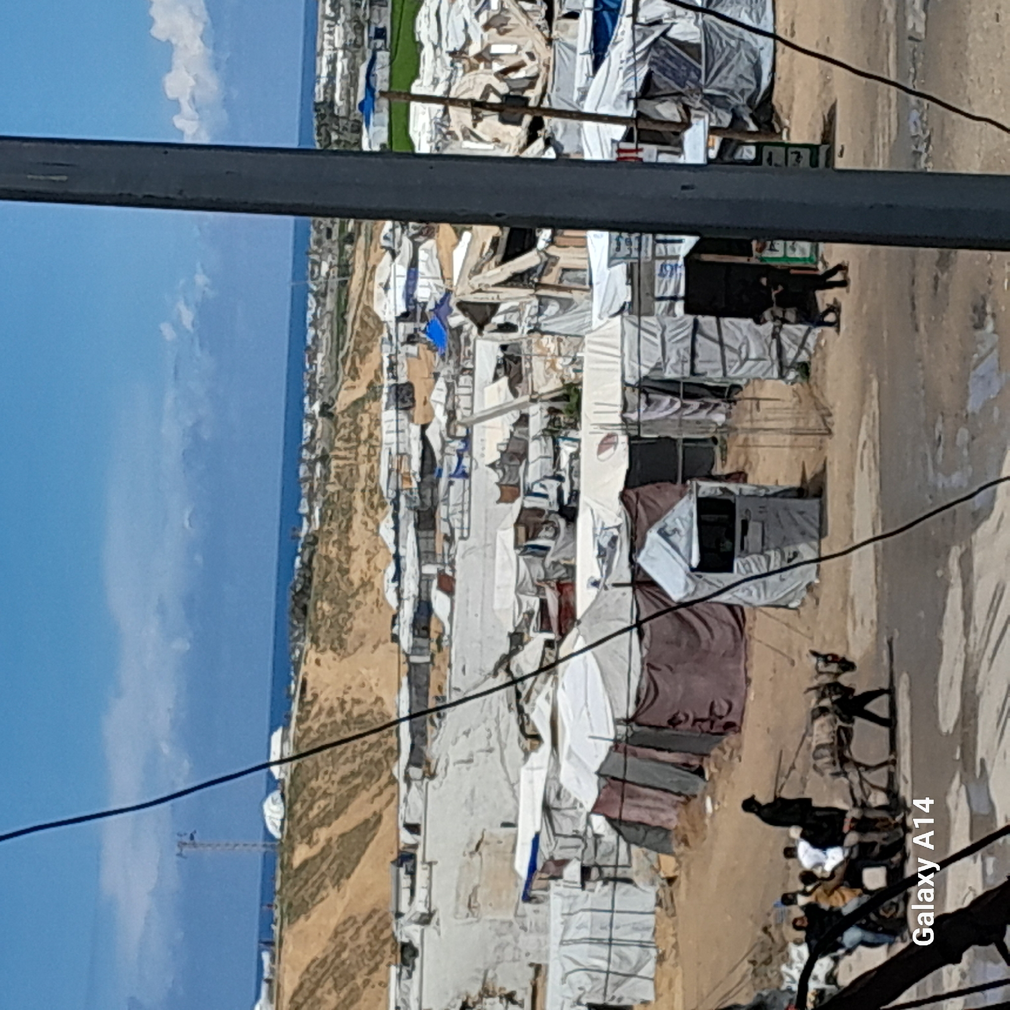 The image shows an overview of a refugee camp, likely located near a coastline. The camp consists of numerous tents and makeshift shelters, densely packed together. People can be seen moving around the camp. In the background, there is a view of the sea under a cloudy sky. Overhead are powerlines and metal structures that appear to be part of the environment, possibly infrastructure elements. The general setting suggests an impoverished and overcrowded living condition.