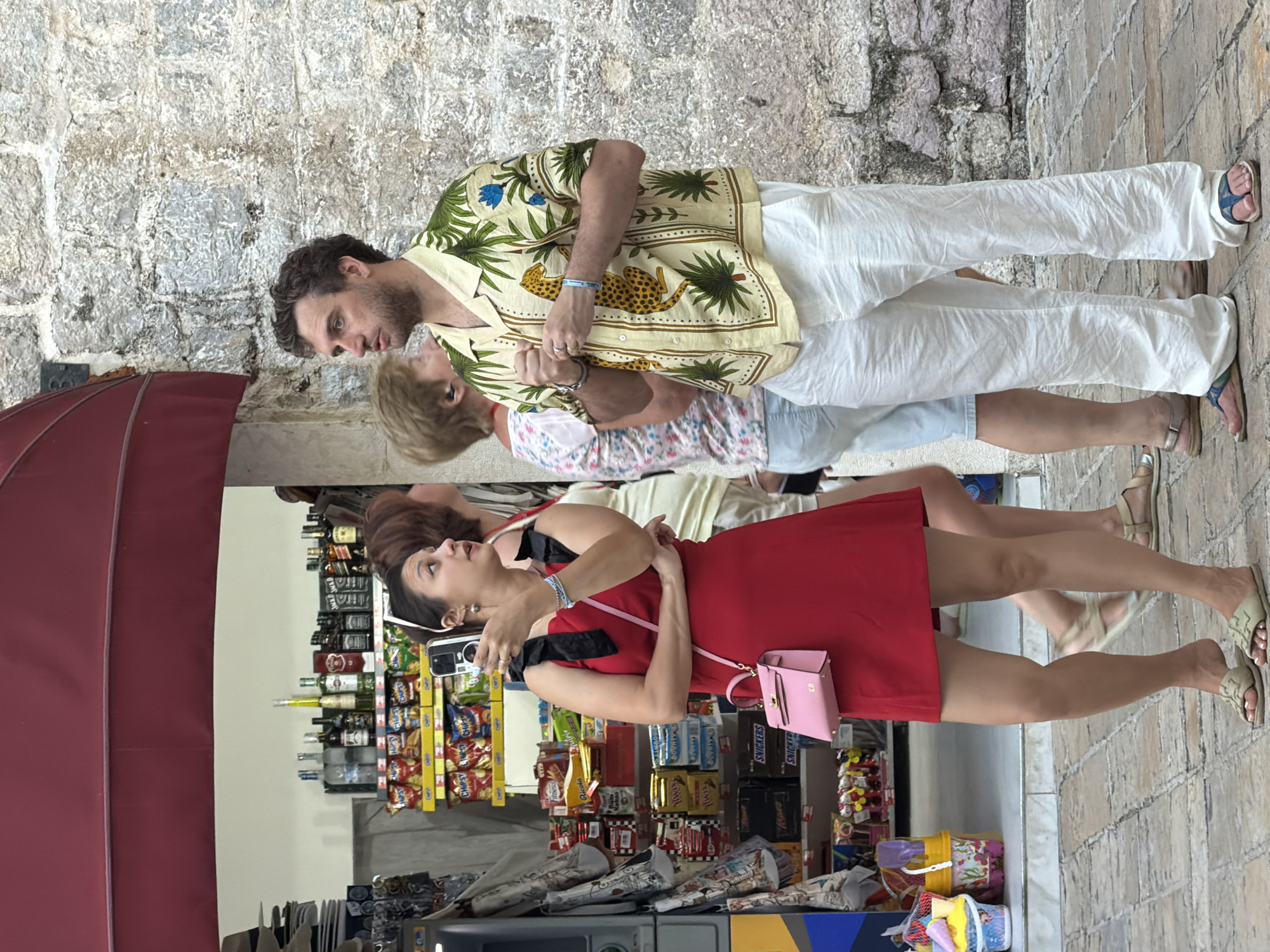 In Budva, Montenegro, a man and a woman stand outdoors on a stone-paved street. The man, wearing a light-colored short-sleeved shirt with a leopard and tropical plant pattern and loose white pants, looks to his left. He has his hands clasped in front of him. The woman, in a red dress with black straps, holds a phone in her right hand and appears to be taking a picture. She wears a small pink handbag with a strap across her body. In the background, a storefront displays shelves stocked with various food items, including chips and candy bars, and bottles of alcohol. The architecture suggests an old town setting, with a stone wall to the right of the man.