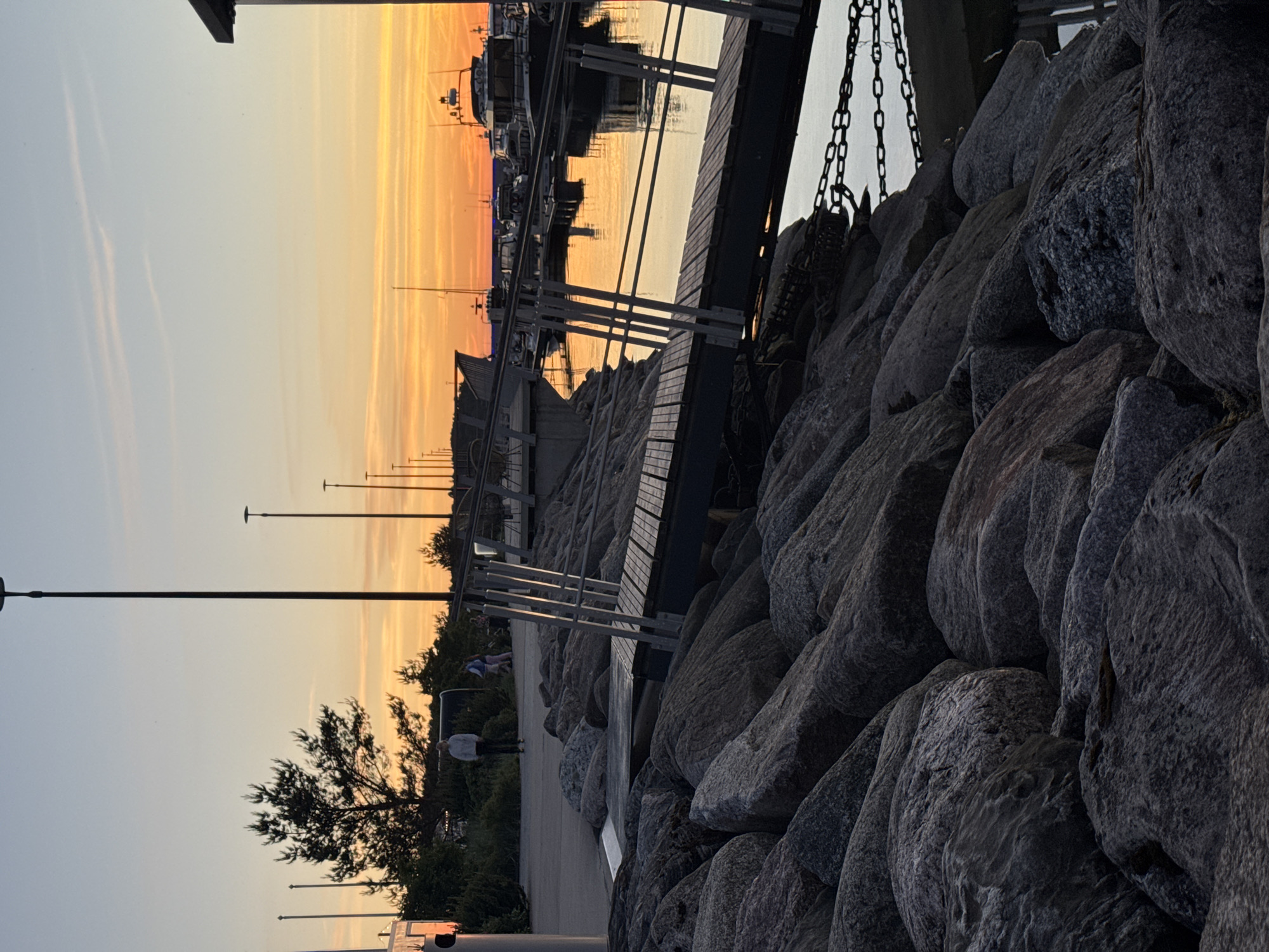 This image depicts a harbor scene at sunset in Tallinn, Estonia. In the foreground, large, grey rocks form a barrier, with a wooden walkway and metal railings extending into the frame from the right. The water in the harbor reflects the warm orange and yellow hues of the sunset. Several boats and yachts are docked in the background. A paved walkway runs along the top of the rocky embankment, where a few people are visible, including one person standing and looking towards the water, and others walking in the distance. Tall lamp posts line the walkway. The sky displays a gradient of soft blue at the top transitioning to vibrant orange and yellow near the horizon. The overall atmosphere is tranquil, characteristic of a harbor at dusk.
