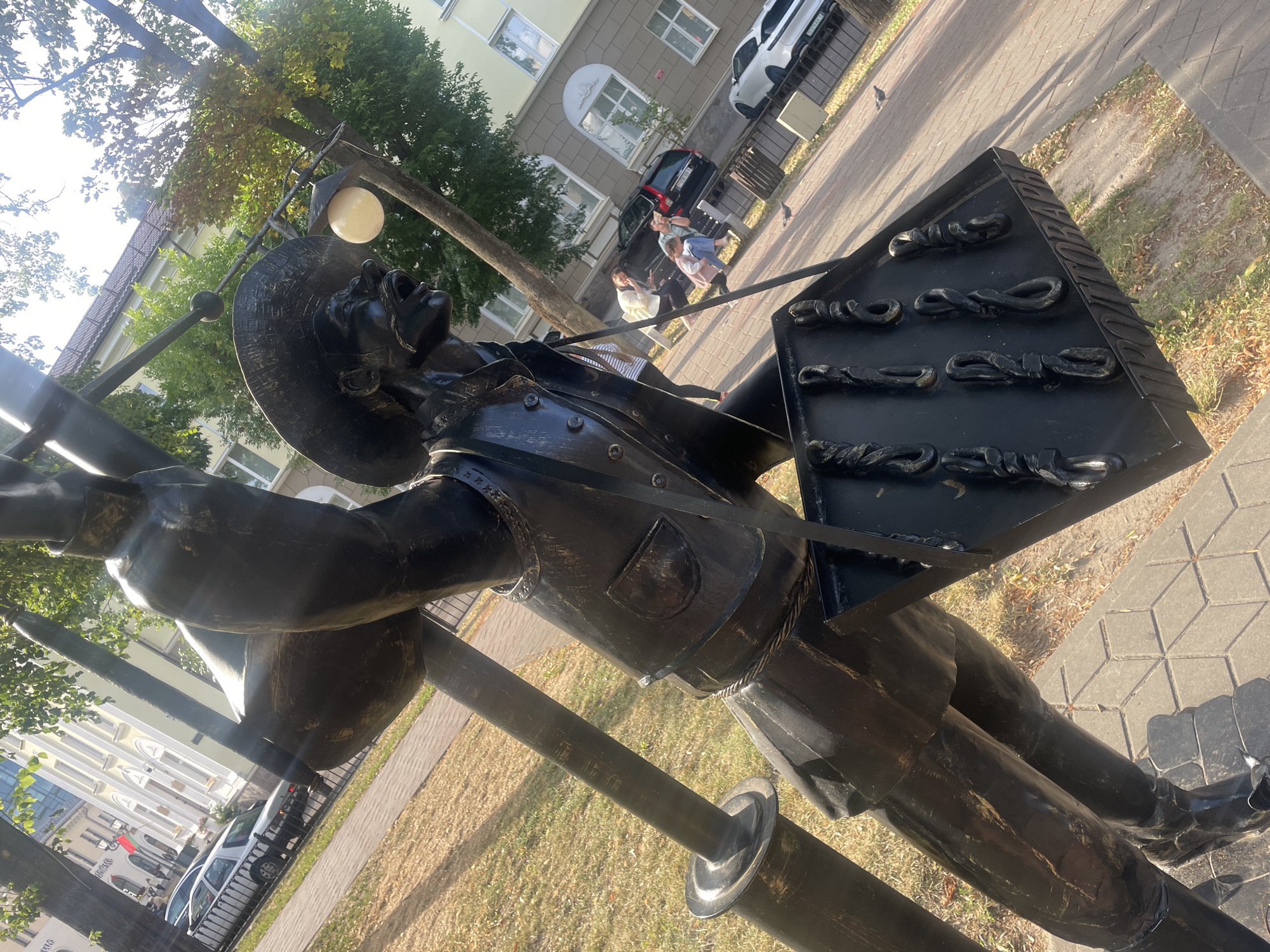 The image shows a dark metal sculpture of a man with an open mouth and wearing a wide-brimmed hat. He appears to be holding a musical instrument, possibly an accordion, and a case or board with several knot-like shapes on it. The sculpture is made of dark metal and is mounted on poles.
In the background, there are several people walking on a sidewalk paved with light-colored bricks. One person is wearing a white jacket and black pants, another is in a pink jacket, and a third is in a light blue shirt. There are also cars parked on the street behind them, including a white sedan and a black SUV. Buildings with windows and balconies are visible behind the cars. Trees with green and yellowing leaves line the sidewalk, suggesting it might be autumn. The sun is shining, casting shadows and creating some glare on the sculpture.
The sculpture is positioned outdoors in what appears to be a public space, likely a street or park. The overall scene suggests a normal, everyday day with people going about their activities. The sculpture itself seems to be a public art piece, possibly depicting a musician or a street performer. There is no visible text in the image.