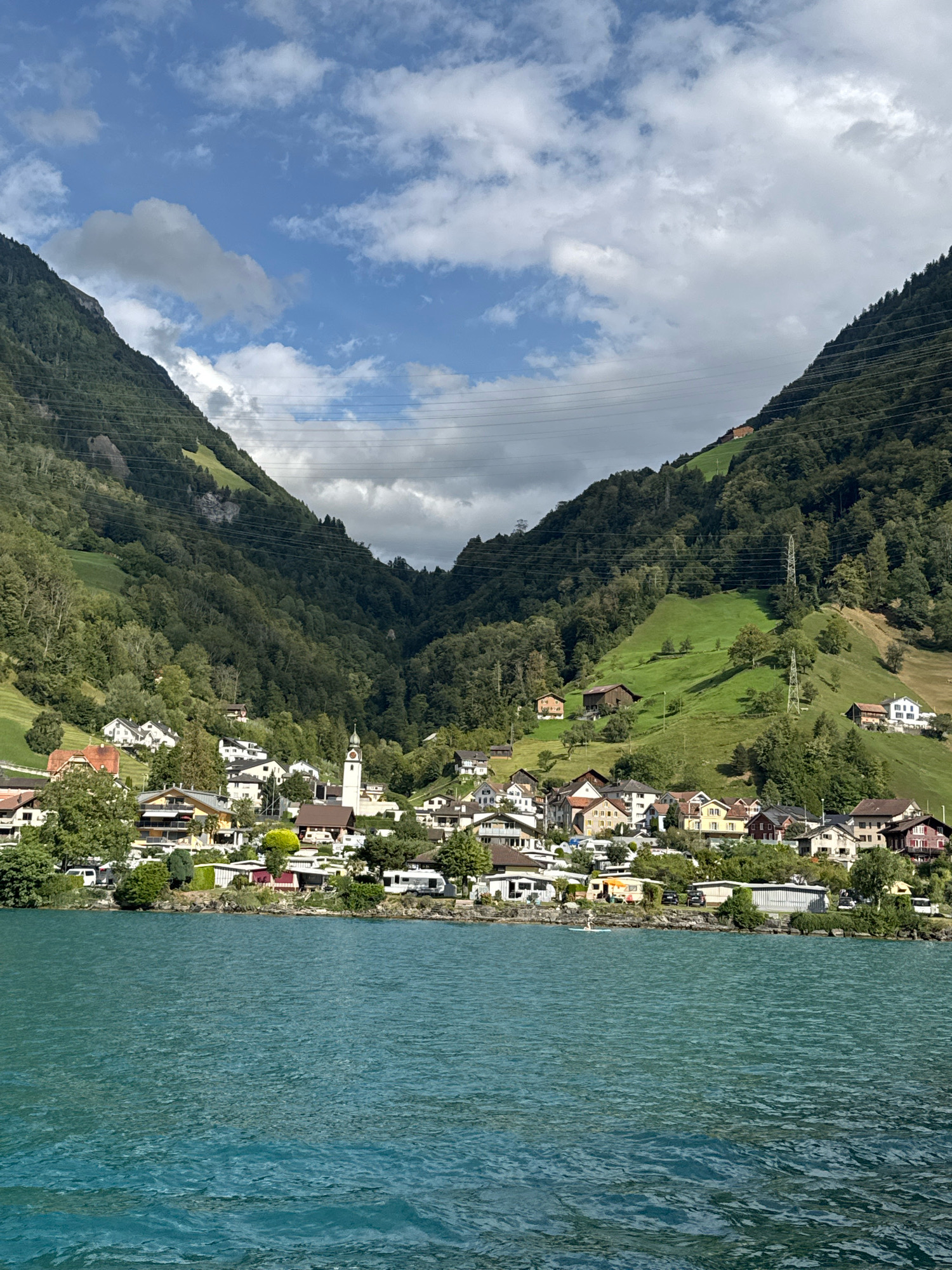 The image depicts a picturesque scene in Sisikon, Switzerland, featuring a village nestled between steep, tree-covered mountains and a vibrant turquoise lake. In the foreground, the calm lake water stretches across the frame, with subtle ripples indicating a gentle breeze. The shoreline is lined with a small collection of buildings, including houses and what appears to be a campground, with RVs and cars visible. A prominent church with a white steeple stands out amongst the residential structures. The midground showcases the village itself, a cluster of multi-story buildings with various colored roofs and facades, situated on the lower slopes of the mountains. Beyond the village, the mountains rise dramatically, their green slopes dotted with patches of exposed rock and traversed by power lines. The sky above is a mix of blue and white clouds, with sunlight illuminating parts of the landscape, creating a sense of a bright, clear day. A single individual can be seen on a paddleboard in the lake, adding a touch of human activity to the serene natural setting.