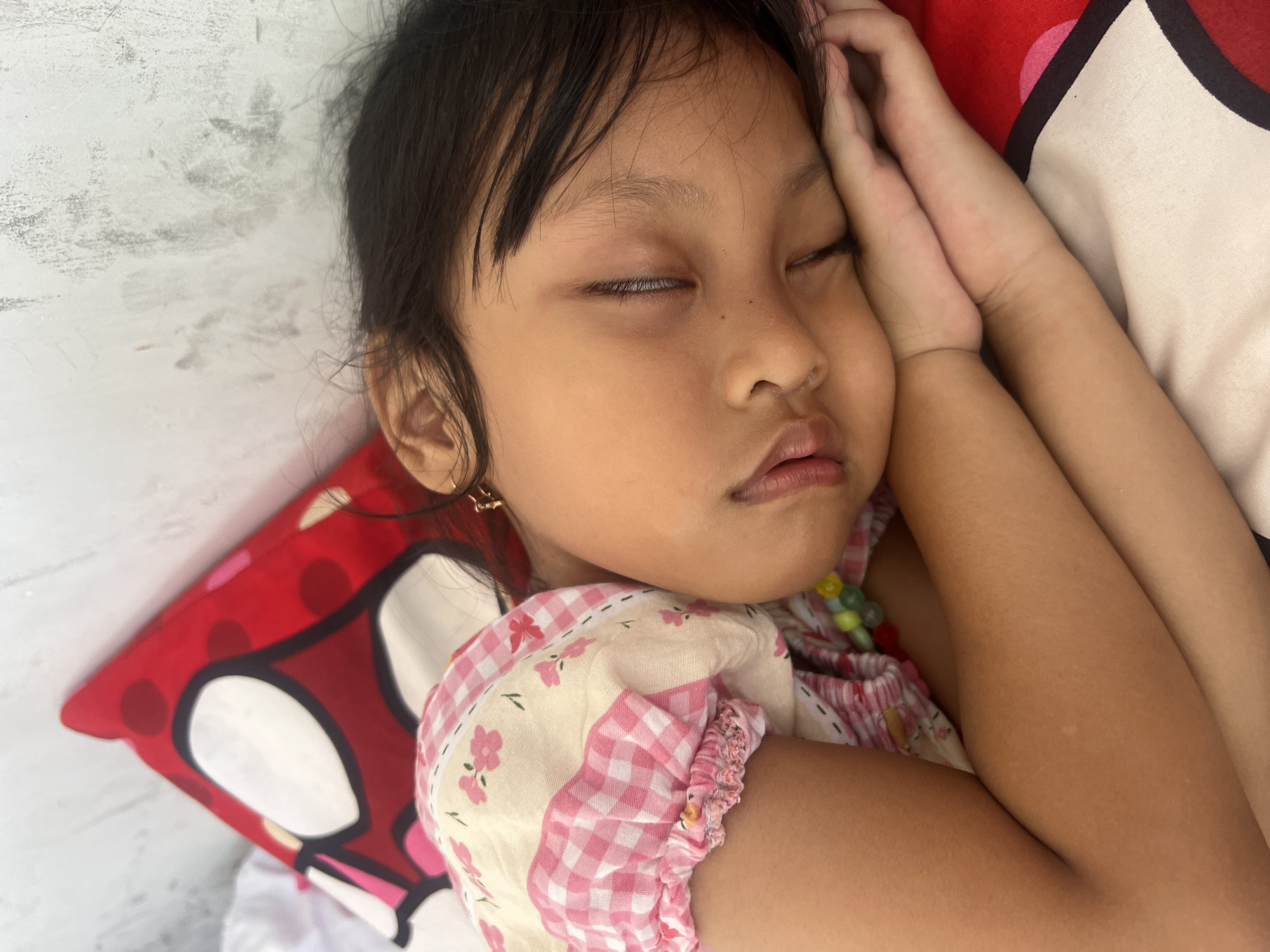 A young girl sleeps soundly, her head resting on her arms, which are positioned beneath her cheek. She is wearing a pink and white checkered dress with a floral pattern. Her eyes are closed, and her lips are slightly parted. A pillow with a red and white polka dot pattern is visible behind her head. The background appears to be a textured wall. The scene suggests a domestic environment, likely a bedroom, and the lighting indicates it is daytime. The image does not provide specific clues to pinpoint the exact location within Kecamatan Pati, Indonesia, but it depicts a personal, intimate moment of sleep.