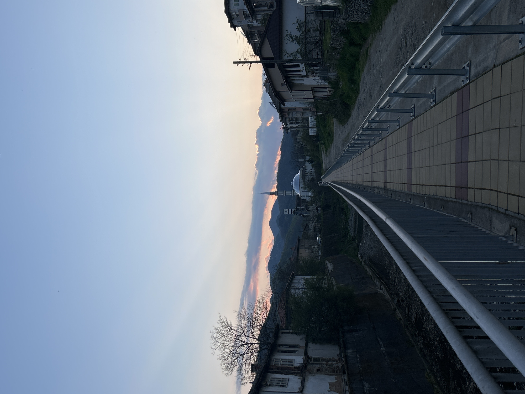 The image shows an upward-angled view of a pedestrian bridge in Zlatograd, Bulgaria, likely taken during the late afternoon or early evening based on the sky's color gradient.
The bridge is constructed with rectangular tiles that form a tiled walkway, bordered by metal guardrails on each side. One side of the bridge faces a street lined with buildings, while the other side appears to overlook a lower area with vegetation and older structures.
In the distance, a mosque with a prominent minaret and dome is visible amidst rolling hills and a backdrop of pastel-colored clouds near the horizon. An aged tree is observed on the left, adding an aged atmosphere. The sky transitions from a pale blue to a lighter shade closer to the horizon.