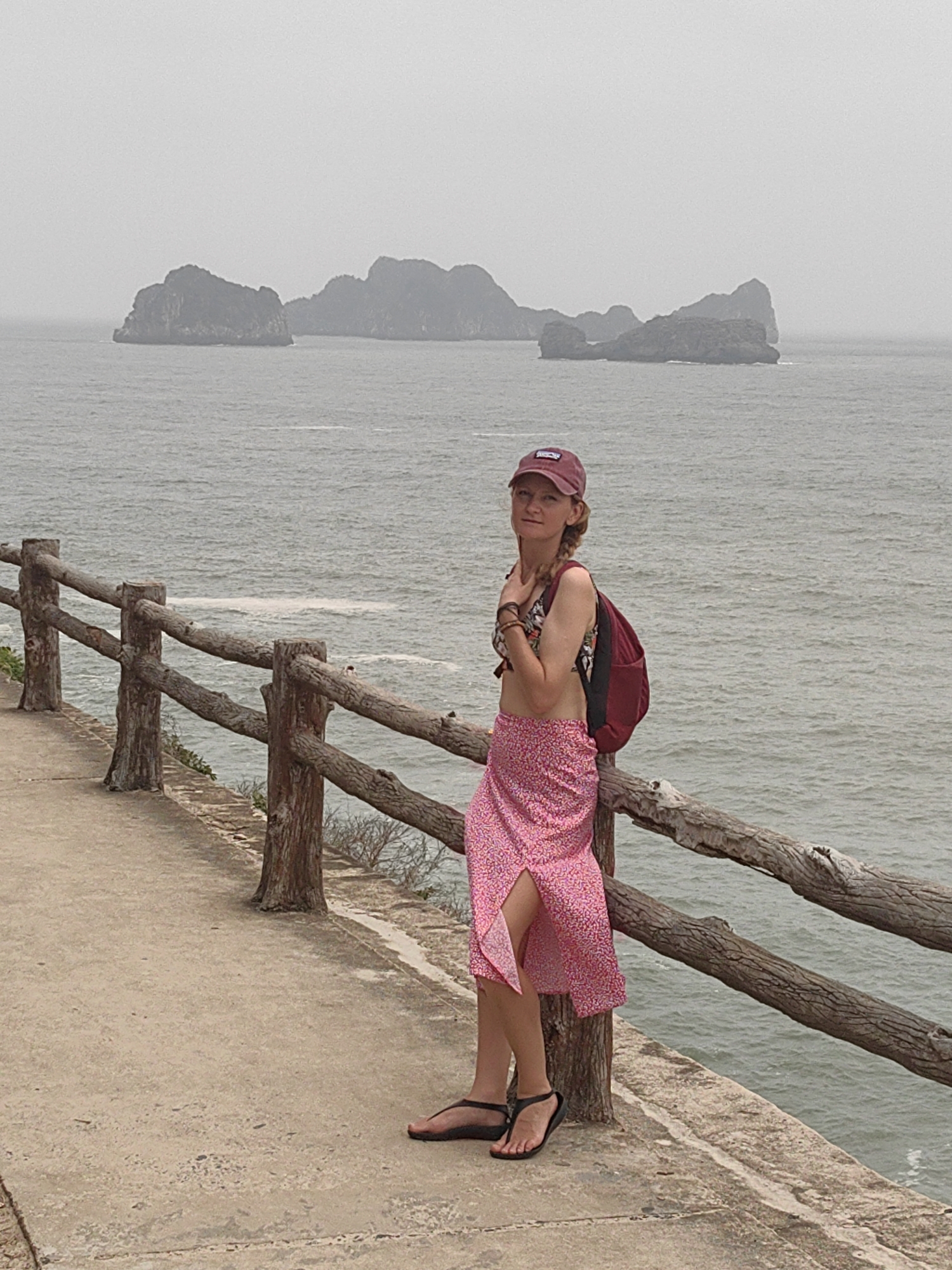A photograph shows a woman standing next to a wooden railing, with the ocean and islands in the background. The woman is wearing a red baseball cap, a floral-patterned bikini top, a red backpack, a pink floral sarong-style skirt, and black sandals. She is leaning against the railing, and appears to be posing for the photo. The islands are in the distance, shrouded in mist. The railing is made of unfinished wood and appears to be a safety barrier along a concrete path. The scene is likely taken on Cat Ba Island, part of the city of Hải Phòng, Vietnam.