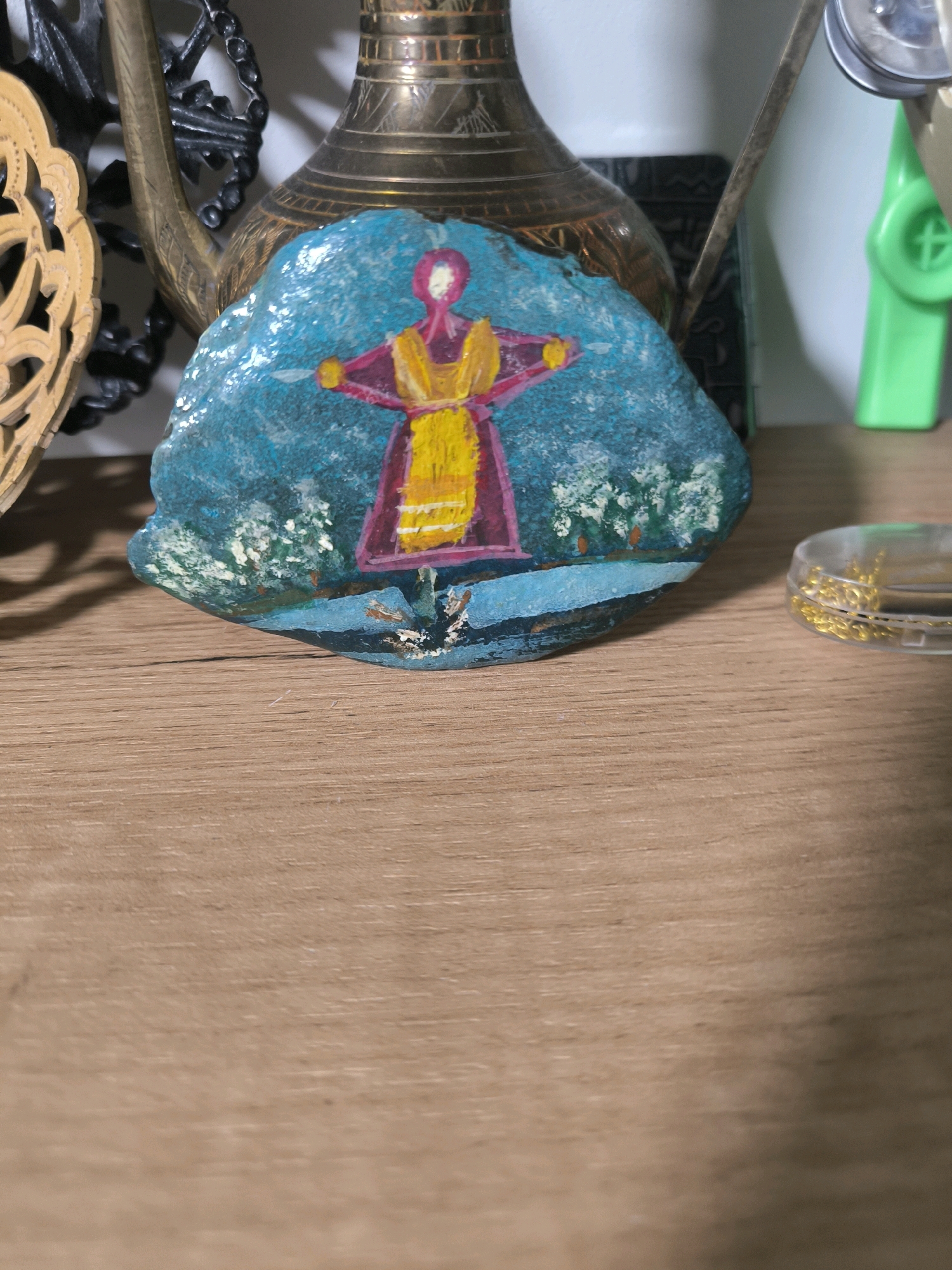 A close-up, eye-level shot displays a painted rock as the central subject, resting on a wooden surface. The rock features a vibrant depiction of a figure in a magenta dress with yellow accents, set against a turquoise background with white and green foliage. Behind the rock, various objects are visible, including a brass vase, a black decorative item, and a small clear container with gold trinkets. The background is blurred, with hints of a green object and a white wall. The lighting suggests an indoor environment. The image does not contain information that would allow for a determination of its location in Magnitogorsk, Russia.