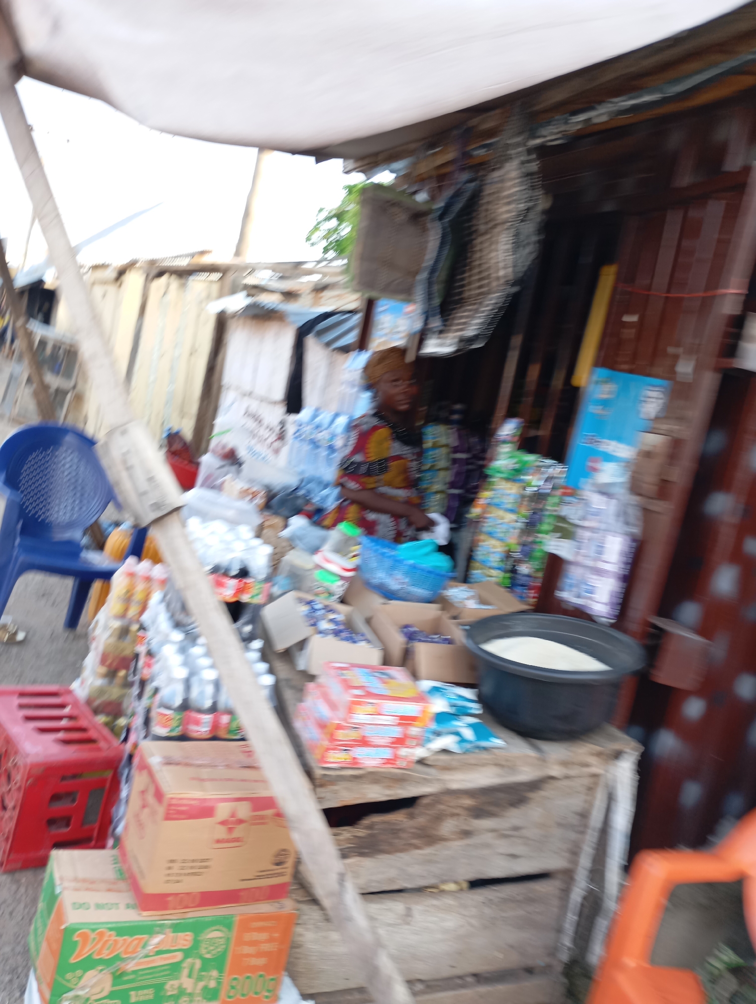 In Shendam, Nigeria, a woman with dark hair wears a colorful patterned top and a headscarf, sitting behind a stall laden with various goods. The stall, constructed from wood and corrugated metal, displays an assortment of items including packaged drinks, boxes of food, and what appears to be rice in a large black basin. To the left, a blue plastic chair is positioned near a red crate. The scene is outdoors, with a light-colored tarp providing partial overhead cover, and wooden structures forming the backdrop.