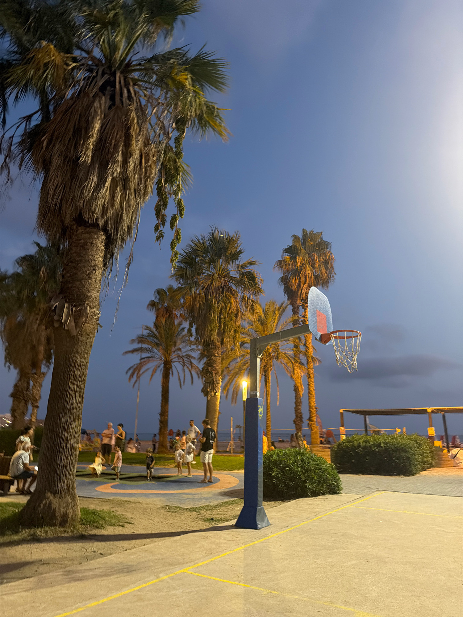 In Salou, Spain, a basketball court with a hoop is visible at dusk. Palm trees surround the court, with some showing dry, hanging fronds. Several people are present in the background, some seated on a bench, while others stand or walk near a playground area. The sky is a deep blue, transitioning to a lighter shade on the right side. A bright light post illuminates part of the scene. In the distance, a beach and structures are discernible. The court itself is paved concrete with yellow lines marking boundaries.