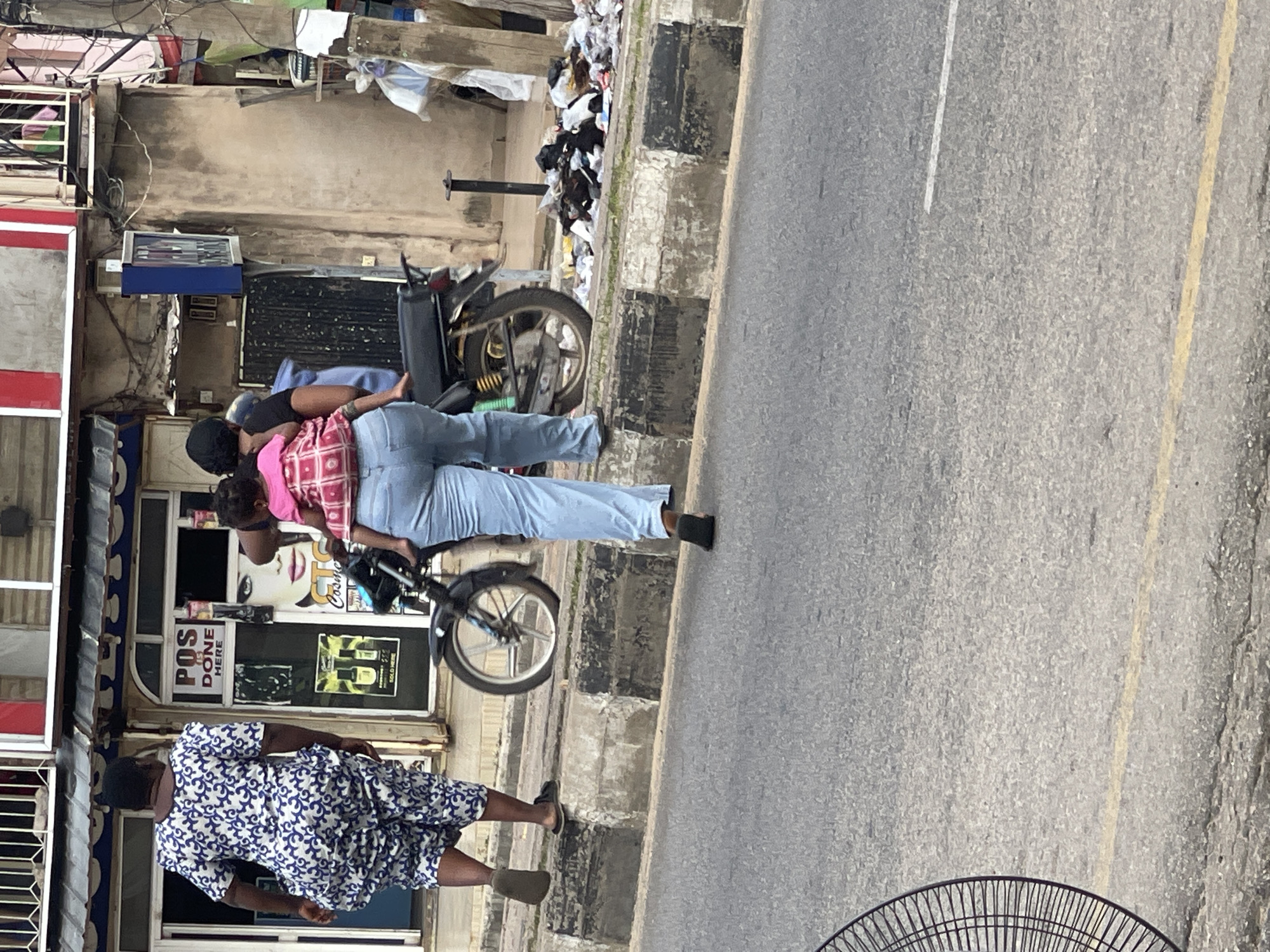 In an outdoor urban scene, a woman in a blue and white patterned dress walks on a sidewalk. She is carrying a child on her back. The child is wearing a pink and white checkered top and blue jeans. Behind them, a motorcycle is parked on the sidewalk. In the background, a building with shops is visible, one of which has a sign that says "POS DONE HERE." There is a pile of trash on the sidewalk near the motorcycle. The ground is paved with asphalt, and there is a curb between the sidewalk and the street. A portion of a metal fan is visible in the lower left corner of the image.