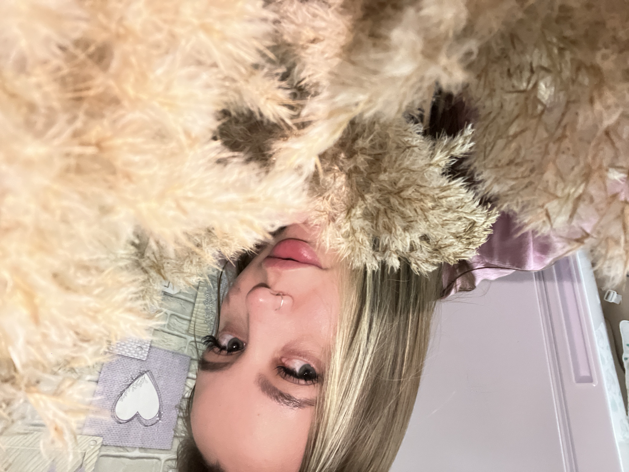 A woman with blonde hair and fair skin is lying down, surrounded by dried plants. She has a nose ring and visible eyelashes. The background includes a lilac-colored surface and a patterned object with a white heart shape on it. It is difficult to determine the specific location in Port-Katon, Russia, based solely on the image content.
