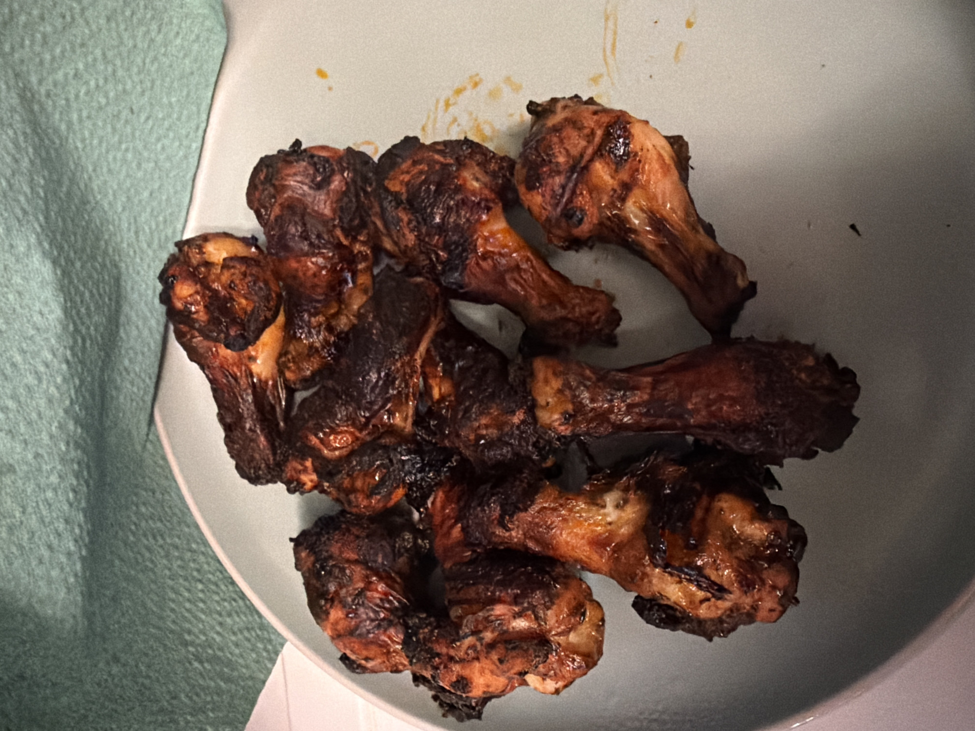 A plate of roasted chicken wings sits on a light blue plate. The wings are dark brown with some charred spots, indicating they are well-cooked. They are arranged in a pile in the center of the plate. A teal-colored fabric is visible in the background. There are no people or any other objects present in the image. The lighting is dim, casting shadows on the plate and wings. The image does not contain any information that would allow for a determination of its location in Bila Tserkva, Ukraine.
