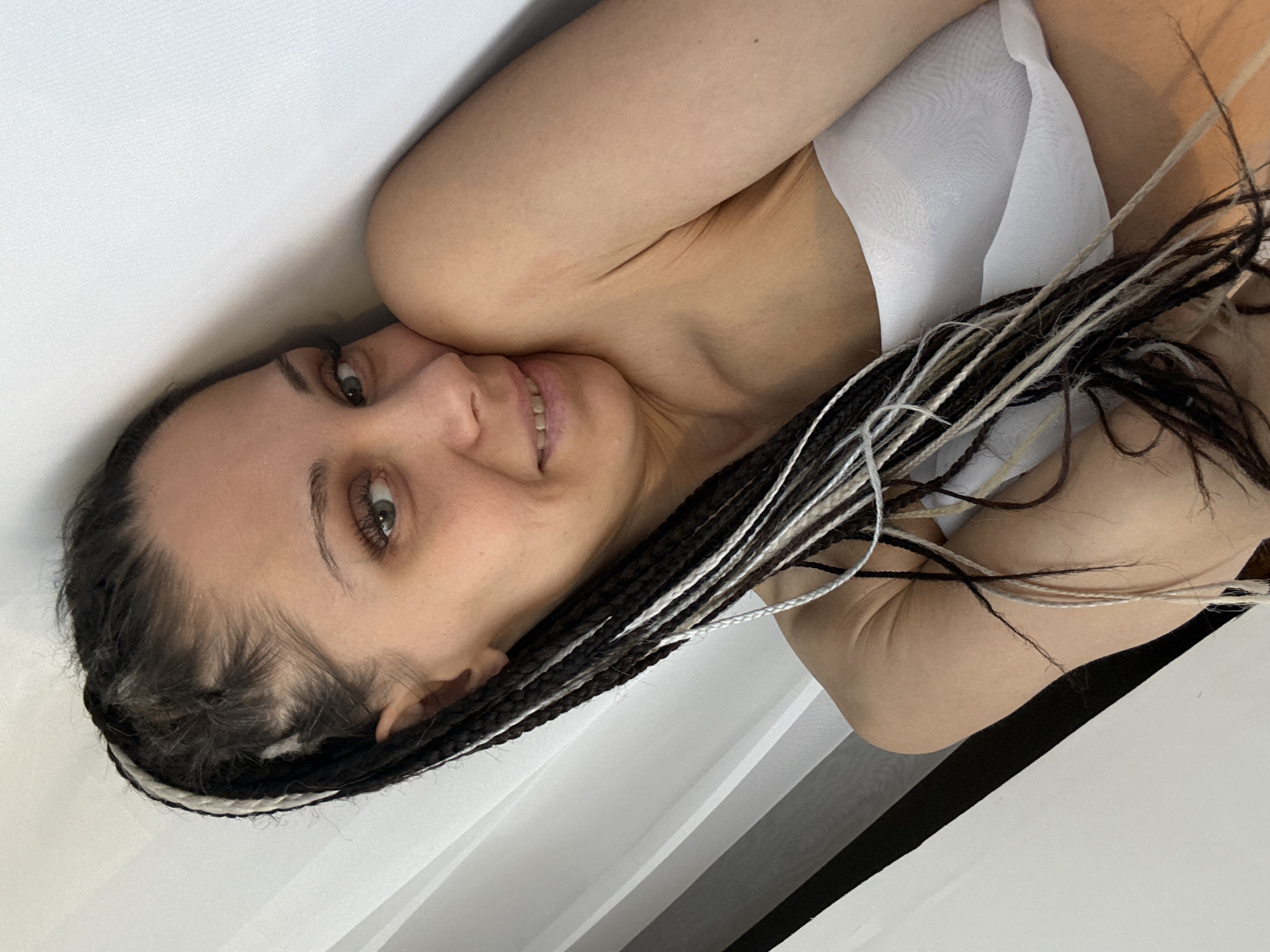 A woman with braided hair, some strands of which are white, lies on a white surface. She is wearing a white top. The background is a blend of white and black elements. There's no indication of the location being Rusanovo, Russia.