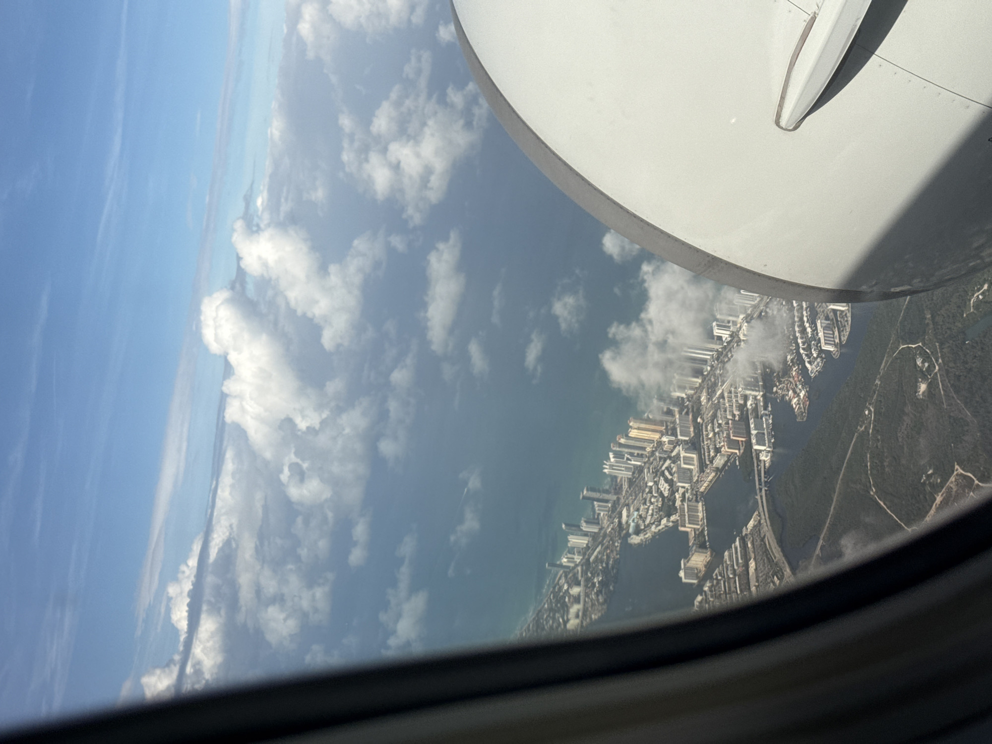 The image shows a view from an airplane window. The wing of the plane is visible in the upper right. Below the wing, there's a partial view of what appears to be the city of North Miami, Florida, USA. The coastline is visible, with high-rise buildings lining the shore. There are also inland waterways and forested areas. Above the coastline, there are scattered clouds in the sky.