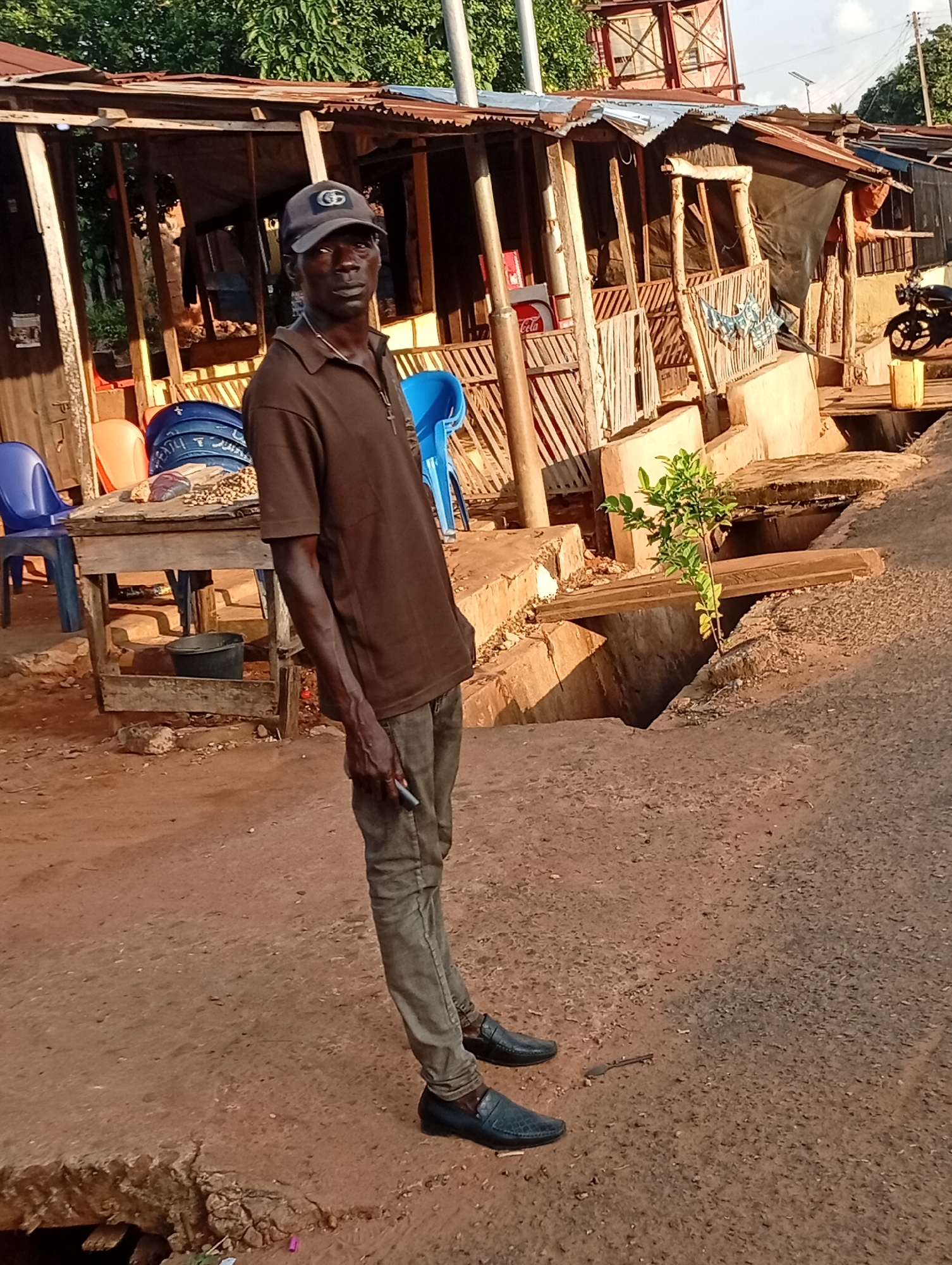A man stands on a dirt road in Nsugpe, Nigeria, in front of a makeshift structure. The man, wearing a dark brown polo shirt, dark pants, a baseball cap, and dark shoes, is looking towards the camera. He holds a small object in his right hand. The structure behind him is made of wood and corrugated metal, with various items visible within, including blue plastic chairs, a wooden table with goods on it, and a black bucket. A small green plant grows in a concrete channel to the right of the man. The ground is a mix of dirt and gravel, with some evidence of erosion and damage. A motorcycle is partially visible in the background. The overall impression is of an outdoor market or informal business area.