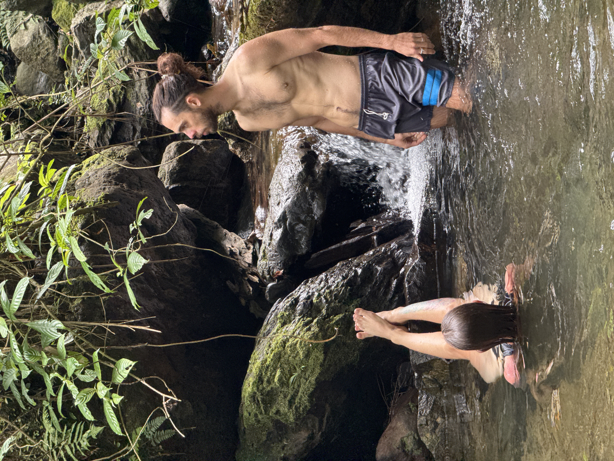A man with long hair pulled into a bun, wearing dark shorts with blue accents, is waist-deep in a stream or river. He leans forward, partially submerged in the water. Opposite him, a person with long hair is face-down in the water, hands extended. The setting appears to be a natural area, likely Suối Giàng, Vietnam, with moss-covered rocks and lush vegetation surrounding the water. The water is clear, with rocks visible beneath the surface.