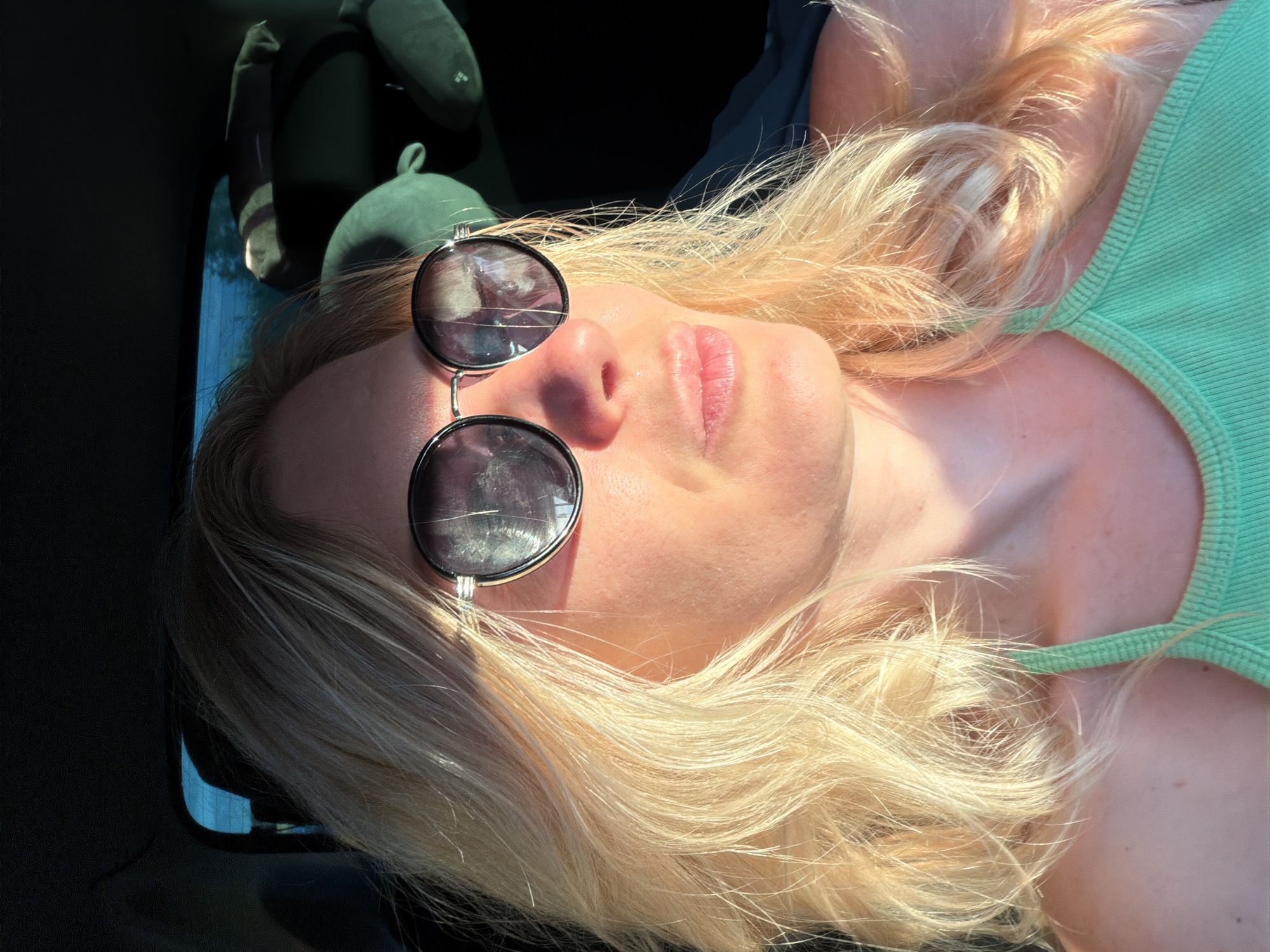 The image features a close-up, overhead shot of a blonde woman lying down, possibly in a car, as suggested by the dark interior and a glimpse of a window. She is wearing a light green tank top and round, dark-lensed sunglasses. The sunlight highlights her hair and face, creating a warm glow. Her face is tilted upwards, and her eyes are not visible due to the sunglasses. The lighting suggests it is daytime, likely with clear skies to produce such direct sunlight. The overall mood appears relaxed and casual, perhaps indicative of a summer day. There is no visible text in the image. The subject's hair is long and light blonde, some strands are catching the light and appear to be blowing slightly. The background is dark and out of focus, with a hint of blue from what could be the sky or a reflection in the window. There's also a dark object with a rounded shape in the upper left corner, possibly a stuffed toy or a headrest.