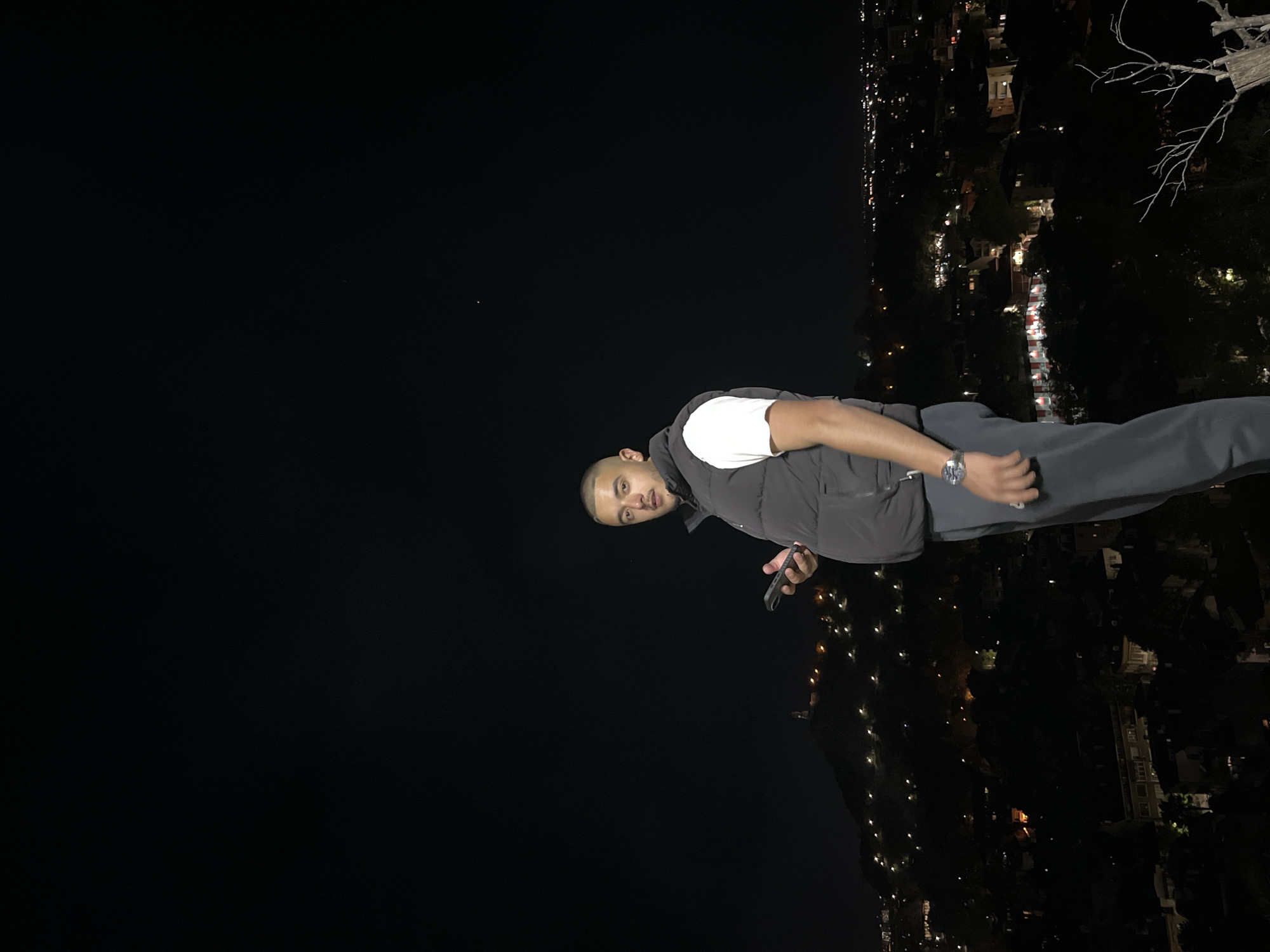 A person stands on an elevated point overlooking a city at night. The person is bald, has tan skin, and wears a white shirt under a dark vest, and dark pants. They hold a smartphone in their left hand and wear a wristwatch on their left wrist. The city below is illuminated with lights, indicating roads, buildings, and possible residential areas. A bare tree branch is visible in the upper right corner. Based on visual cues and available data, the city is likely Plovdiv, Bulgaria, seen from one of the hills overlooking the urban area.