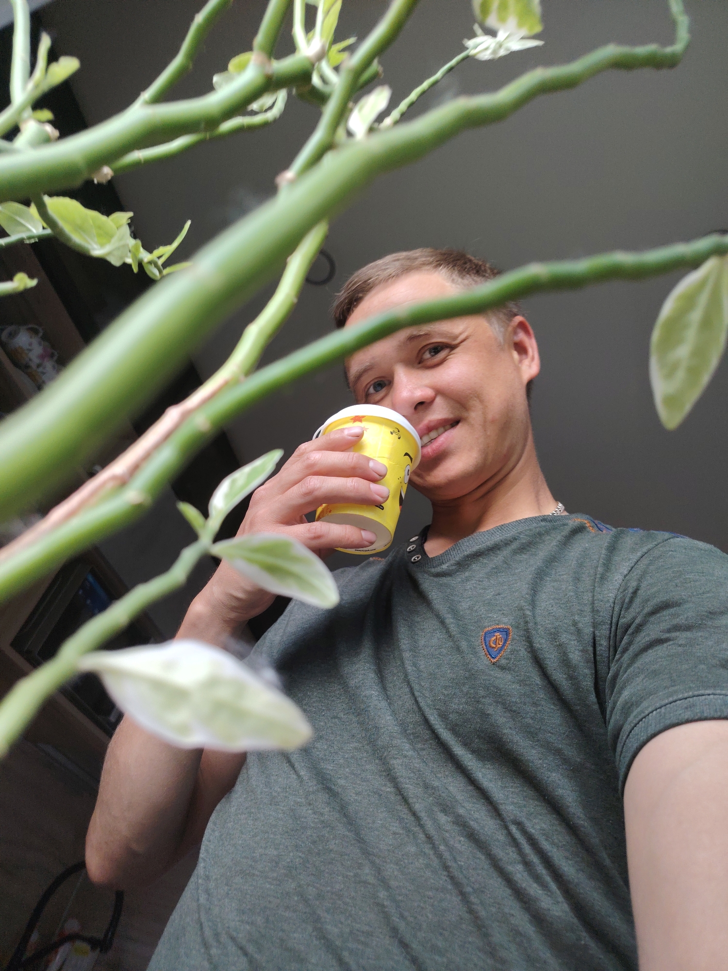 The media file is a low-angle, medium shot of a man holding a yellow cup with a red star design to his lips. He is smiling and looking at the camera. He is wearing a dark green t-shirt with a crest on the left side of his chest. The man is partially obscured by green plant stems and leaves, which are in the foreground and out of focus. The background appears to be an indoor setting with some shelves visible on the left. The overall mood is casual and relaxed. The time of day and weather are not discernible. The location is Chaikovsky, Russia. There is visible text on the cup, but it is not legible.