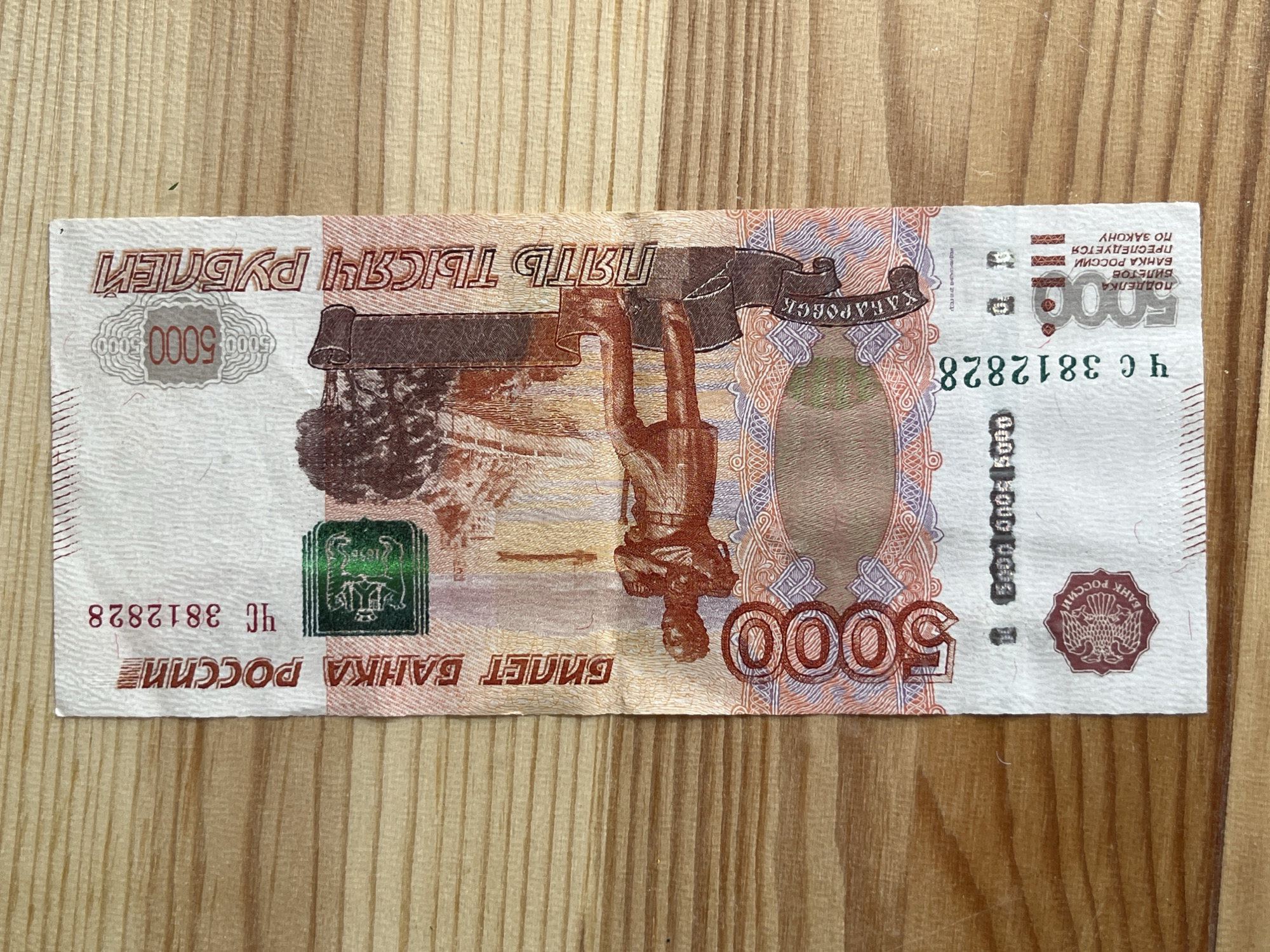 This media file shows a five thousand ruble banknote laid flat on a wooden surface. The banknote is oriented horizontally. The front of the banknote is visible, displaying intricate designs and text in Russian. Prominently featured are the numbers "5000" in large font, along with text indicating "ПЯТЬ ТЫСЯЧ РУБЛЕЙ" (Five Thousand Rubles). There is also an image of a monument and a double-headed eagle emblem. The banknote appears to be in good condition. The wooden surface has a visible grain pattern, suggesting it is a table or countertop. The lighting in the image is even, indicating it was likely taken indoors. There are no people or animals visible in the frame, and the focus is solely on the banknote and its immediate surroundings. The scene is static, with no activity taking place. The text on the banknote includes serial numbers, such as "ЧС 3812828," and other inscriptions related to the Bank of Russia and legal warnings about counterfeiting.