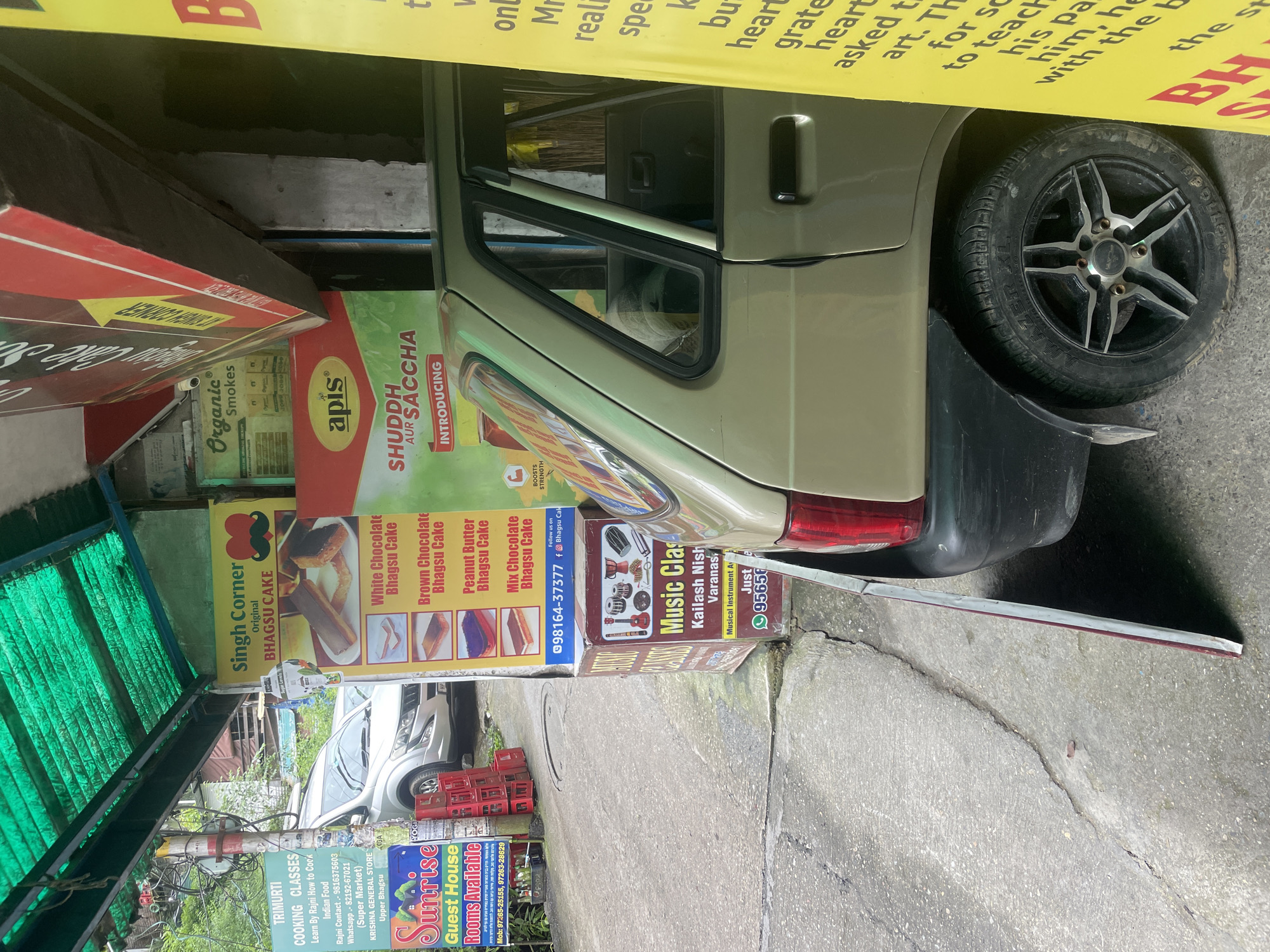 The image shows a street scene in Bhagsu, India, with a car parked on the side of the road. To the left of the car is a green sign advertising "TRIMURTI COOKING CLASSES" and "Sunrise Guest House Rooms Available." Further left, a white SUV is partially visible. To the right of the car, a yellow sign with red text is partially obscured by the car's door. Behind the car, a red and yellow sign for "Singh Corner Original BHAGSU CAKE" displays images of cakes and lists prices for different flavors. Below this, a smaller sign advertising "Music Classes" with images of musical instruments is visible. The environment appears to be an outdoor marketplace or street, with a covered awning overhead.