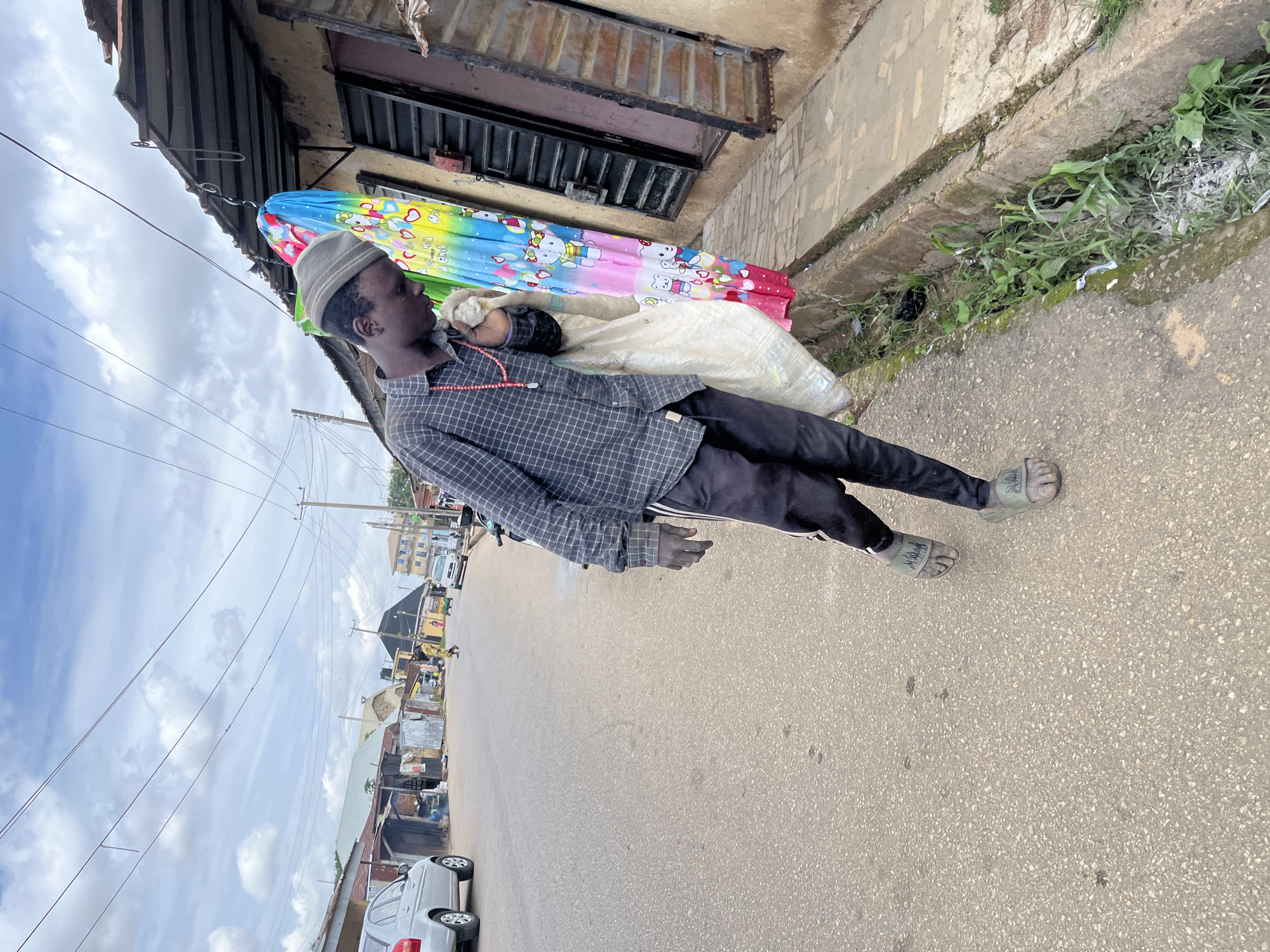 A young man wearing a grey patterned shirt, black pants, and green sandals is walking along a paved road in Jos, Nigeria. He is carrying a large white bag over his right shoulder and has a string of red beads around his neck. In the background, there are several buildings with corrugated metal roofs and a colorful umbrella adorned with cartoon characters. Cars and a motorcycle are visible on the road, and utility poles with wires stretch across the sky. The weather appears overcast with scattered clouds. A concrete drainage ditch runs alongside the road, filled with weeds and some debris.