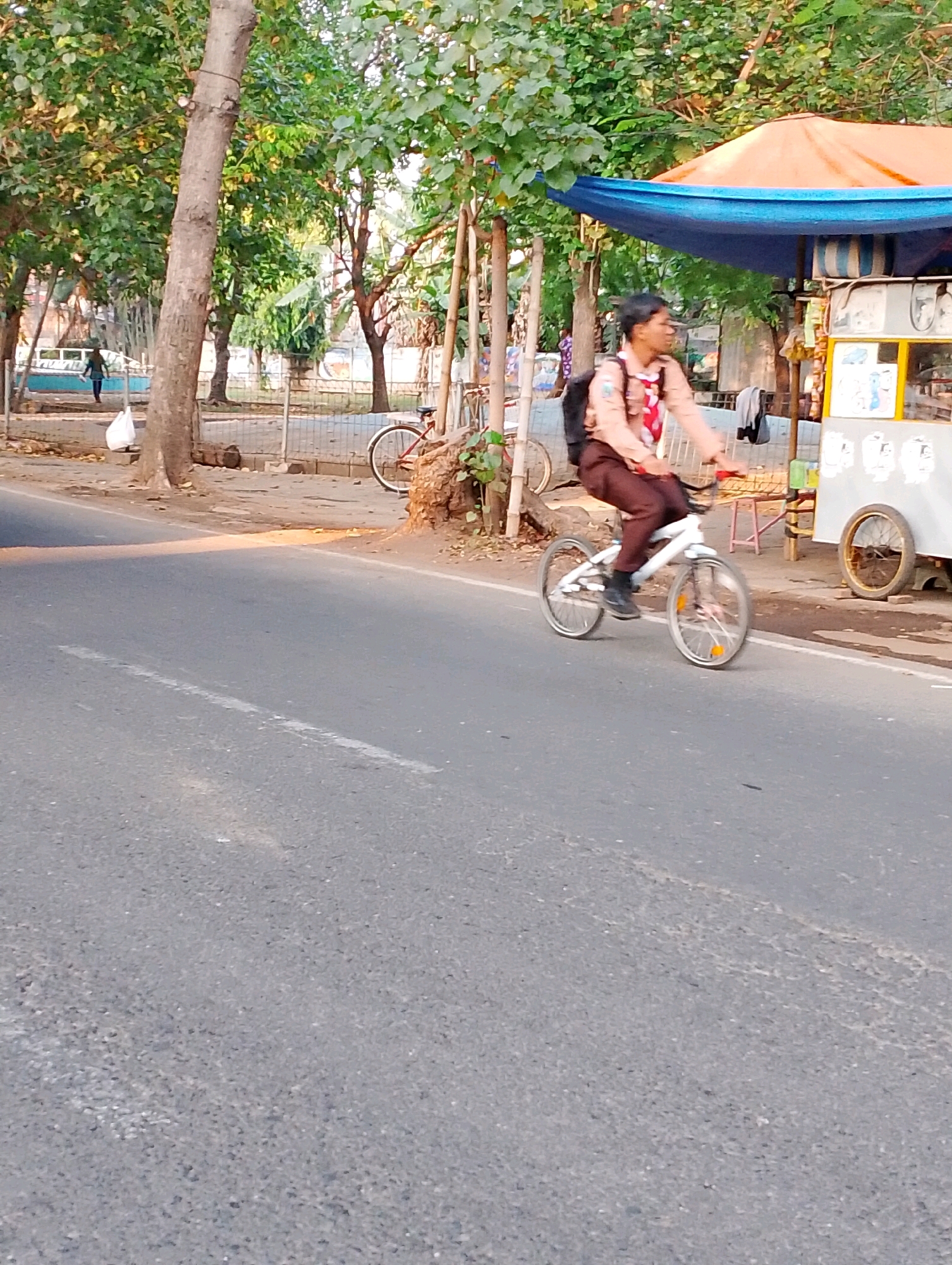 A young person wearing a scout uniform rides a white bicycle on a paved road in Probolinggo, Indonesia. The scout uniform consists of a light brown shirt with a red and white neckerchief and dark brown pants. The person has a black backpack. The bicycle has thin wheels and appears to be a BMX-style bike. They are riding on the right side of the road, with trees lining the left side of the pavement. Behind the scout, there is a food stall with an orange and blue awning, displaying various items for sale. The weather appears to be sunny, with shadows cast on the road. In the background, there are more trees and some fencing. The road surface is asphalt, with a white line marking a lane.
