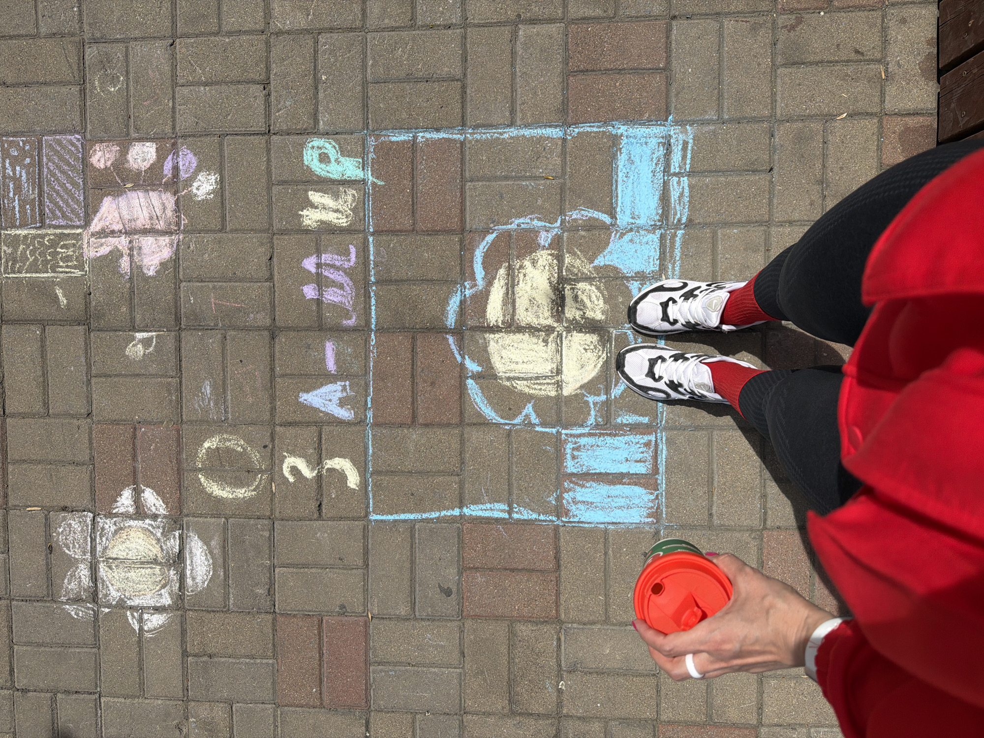A person is standing on a hopscotch game drawn in chalk on a brick-paved surface. The person is wearing white sneakers with black detailing, red socks, and black leggings. They are also wearing a red sweater or jacket. In their right hand, they hold a red and green cup. The hopscotch game is drawn with colorful chalk and includes shapes and numbers. The surroundings include other chalk drawings of flowers and patterns. The location in Birobidzhan, Russia is not discernible from the image.