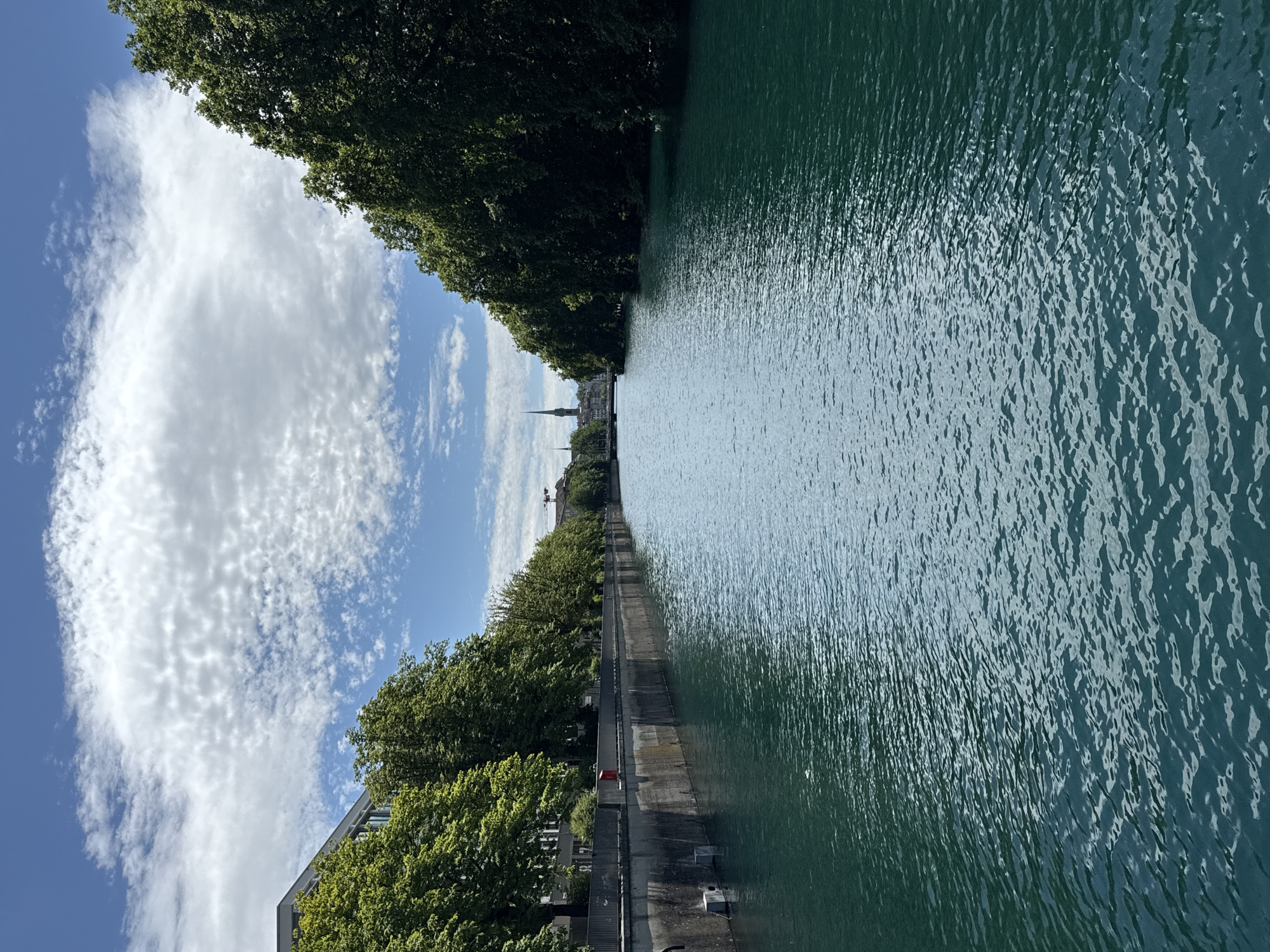 The image depicts a tranquil scene along a river in Zurich, Switzerland. The river, with its characteristic turquoise water, flows through a cityscape lined with lush green trees on both banks. The left bank features a modern building complex and a concrete embankment with a metal railing. People can be seen walking along the embankment. The right bank is densely covered with mature trees, creating a natural buffer between the river and the city beyond. In the distance, the spires of Zurich's churches are visible, a hallmark of the city's skyline. The sky above is a bright blue, dotted with fluffy white clouds, suggesting fair weather. The overall impression is one of urban harmony, where nature and architecture coexist peacefully.
