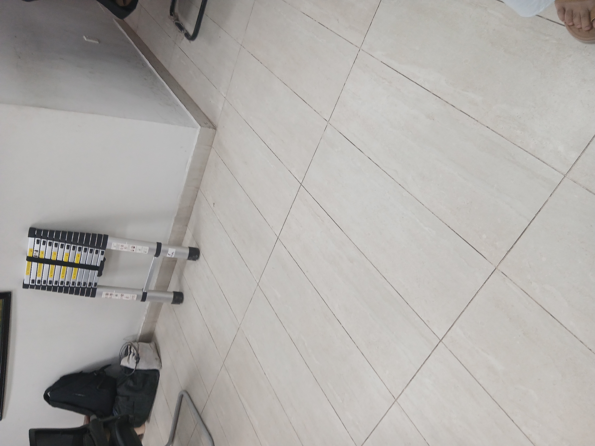 The image displays a sparsely furnished room with tiled flooring. In the background, against a white wall, a black backpack and a grey duffel bag are placed near a chair. To the right of the bags, a silver, foldable ladder is propped against the wall. The ladder is extended in a partially opened position. The environment appears to be an indoor space, possibly an office or a storage area, given the presence of office furniture and storage items. The lighting suggests it is daytime. No individuals are prominently featured in the scene, although a foot is partially visible in the lower right corner. The location within Peshawar City Tehsil, Pakistan is not discernible from the visual information present in the image.